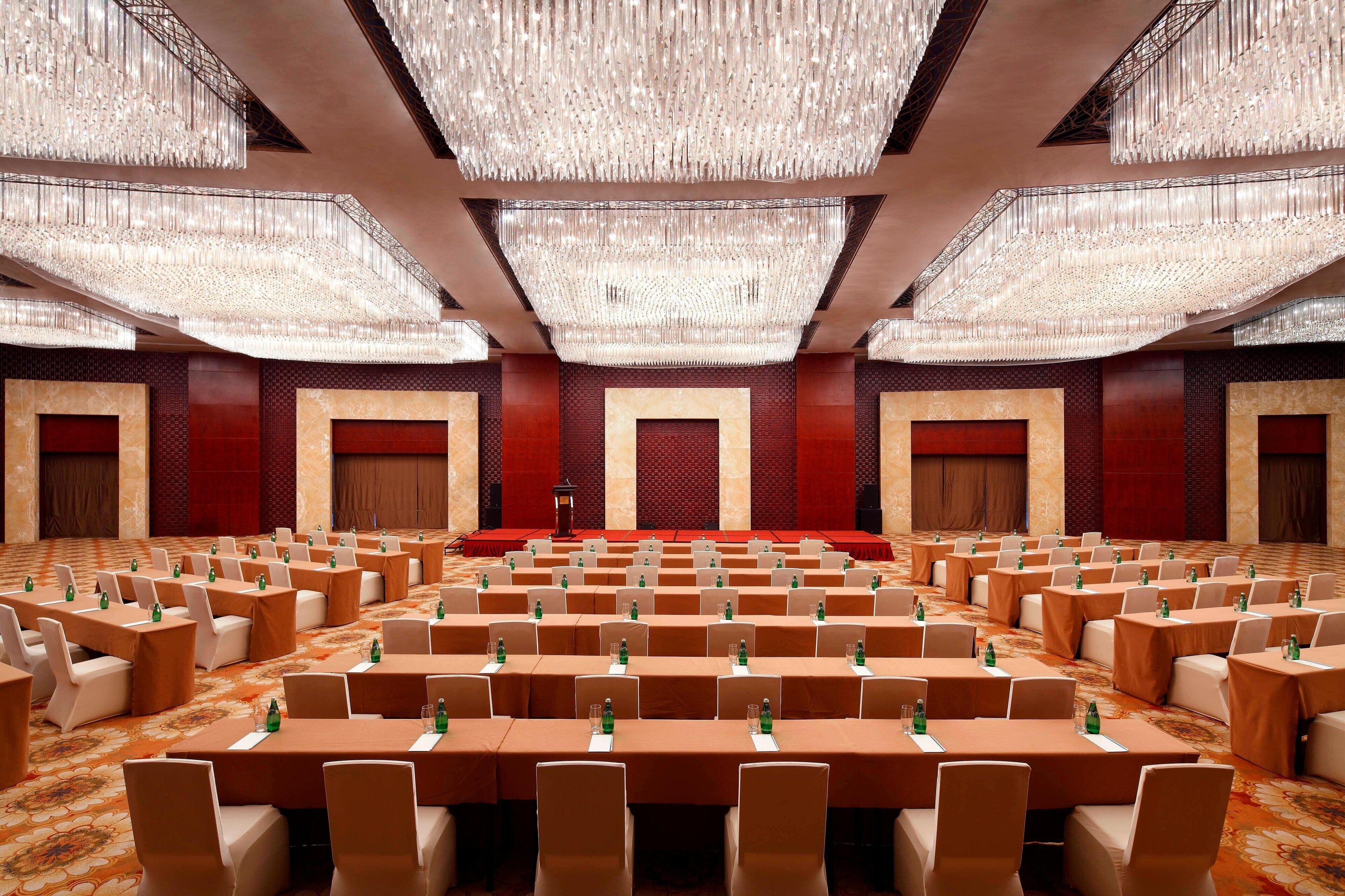 Un amplio salón de conferencias con filas de mesas y sillas, un escenario y candelabros ornamentados.
