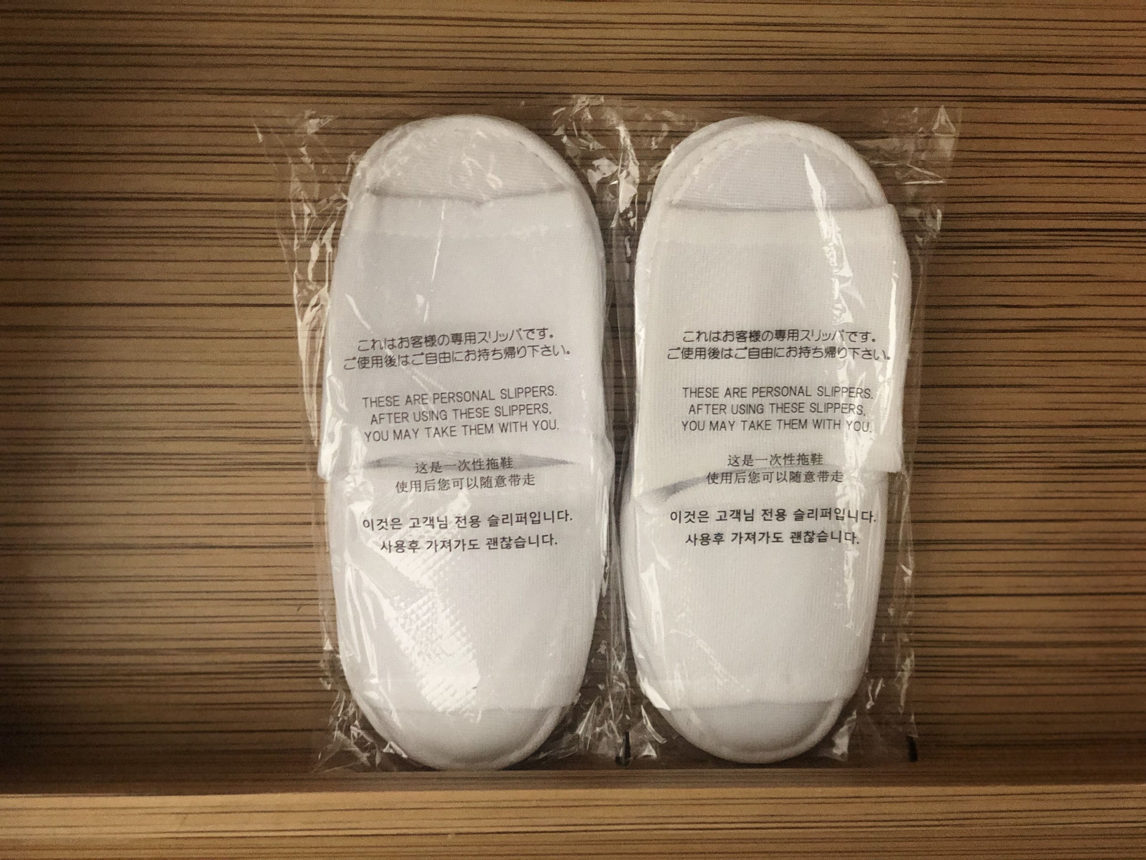 Two pairs of slippers in plastic packaging on a wooden surface.
