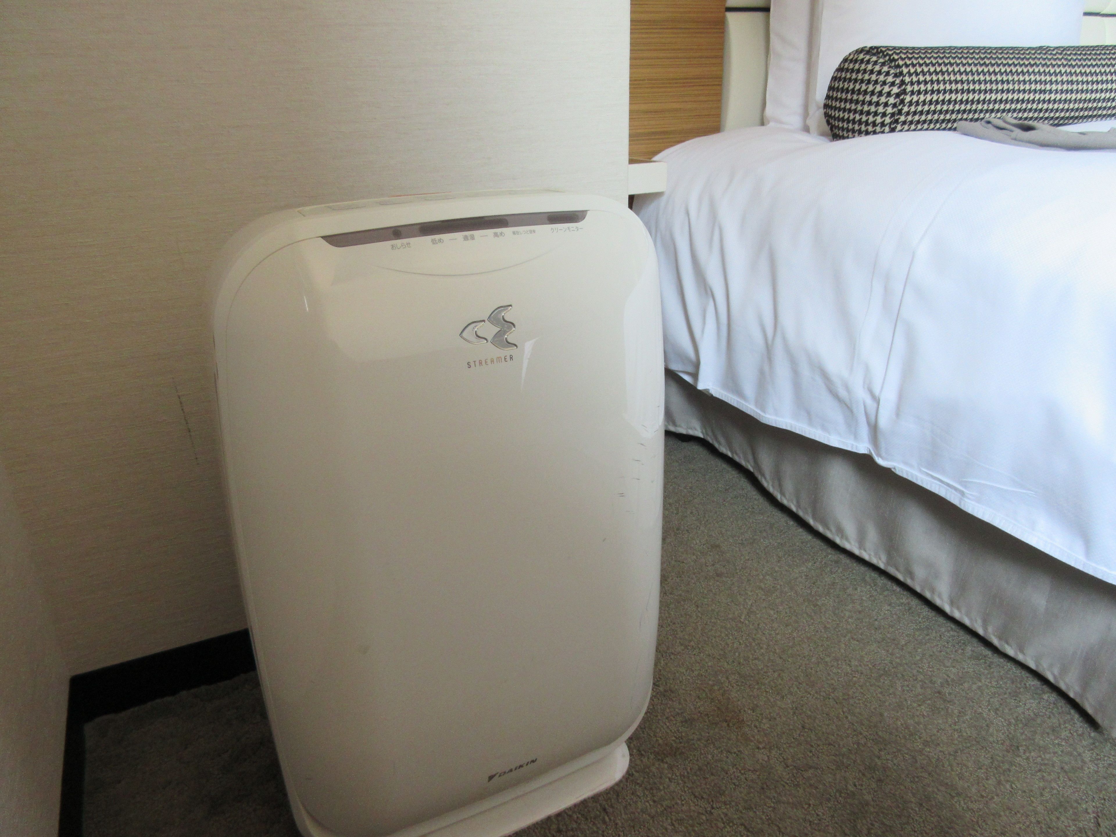 A hotel room with a white bed, a houndstooth pillow, and a white portable heater with the brand name "Streamer" on it.