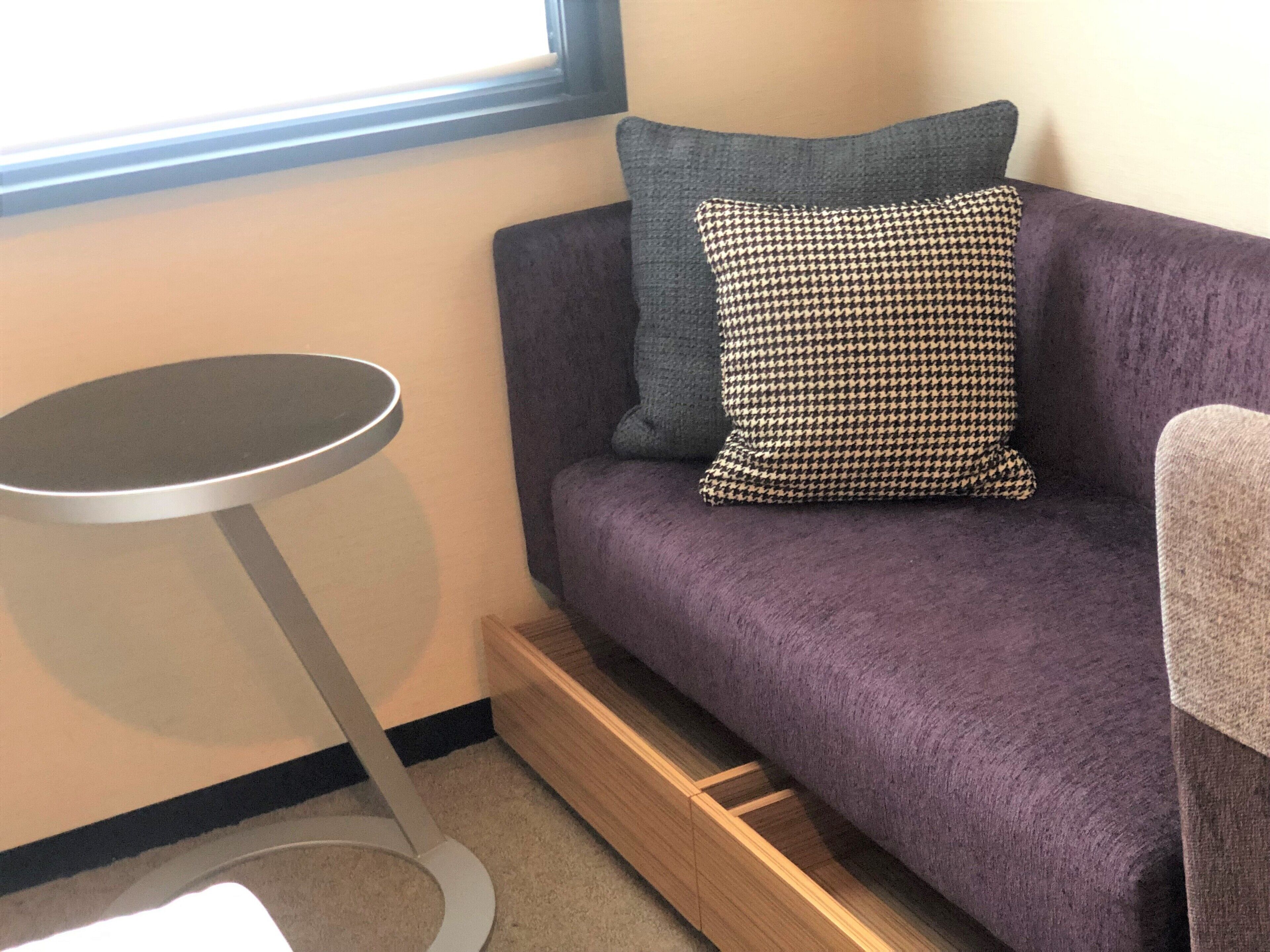 Une pièce avec un canapé violet, un coussin à carreaux, un coussin gris, une petite table ronde et un plancher en bois.