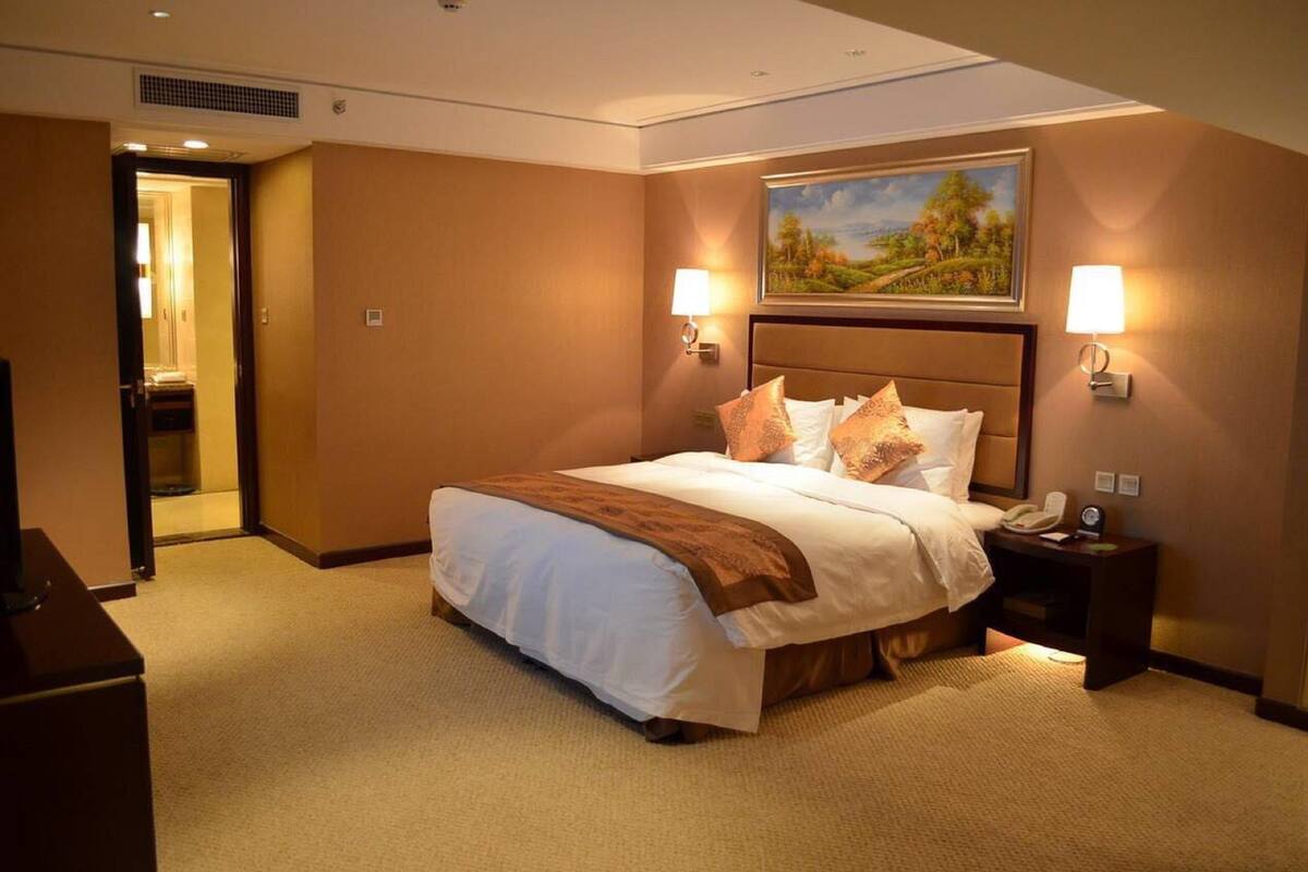 A hotel room with a large bed, two bedside lamps, a painting on the wall, and a door leading to another room.