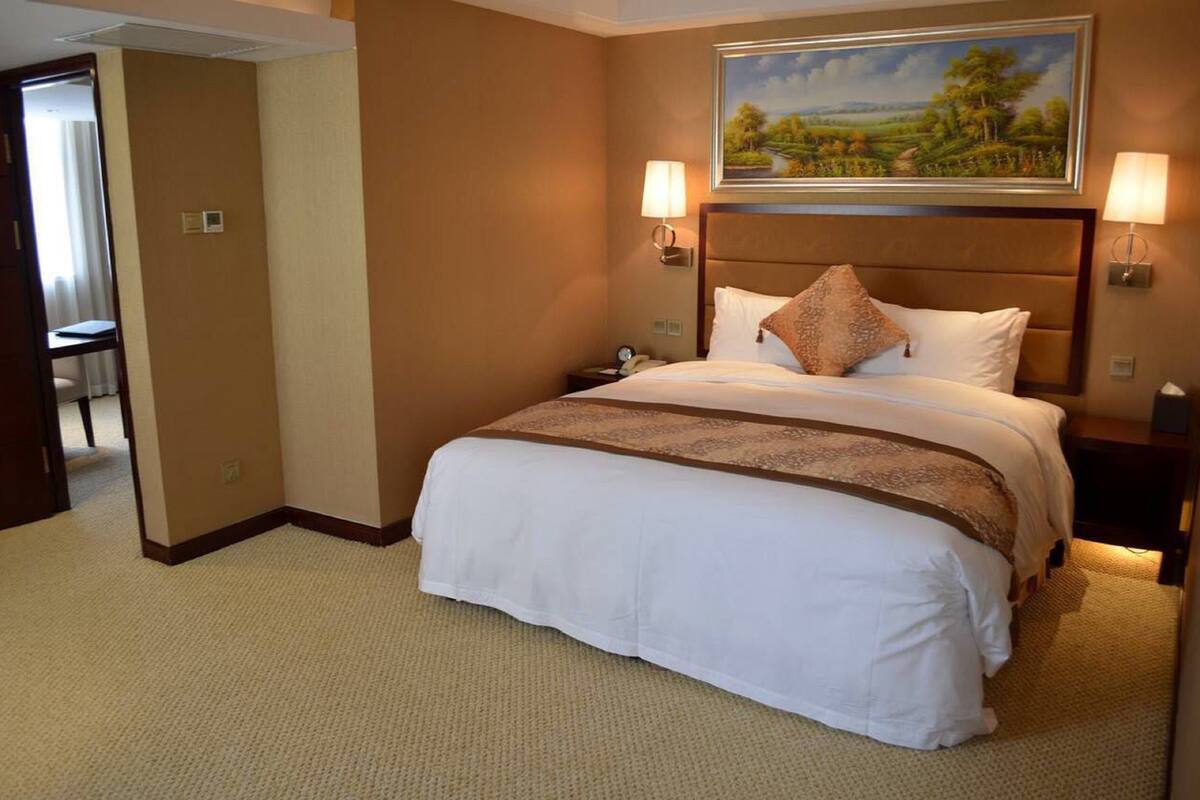 A hotel room with a large bed, two bedside lamps, a painting above the bed, and a door leading to a bathroom.