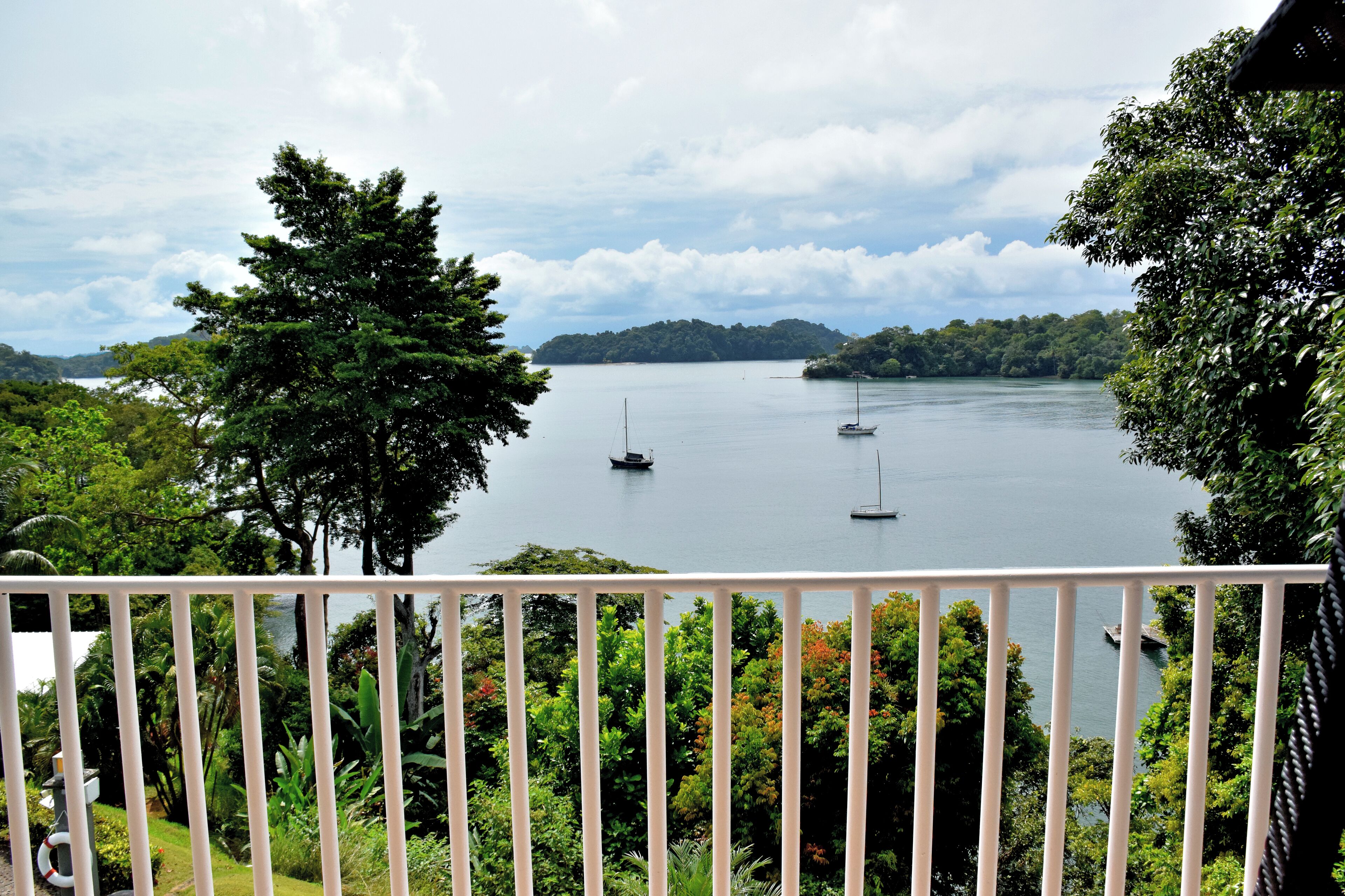 A view from a balcony with a railing, overlooking a calm lake with boats, surrounded by lush greenery and trees.