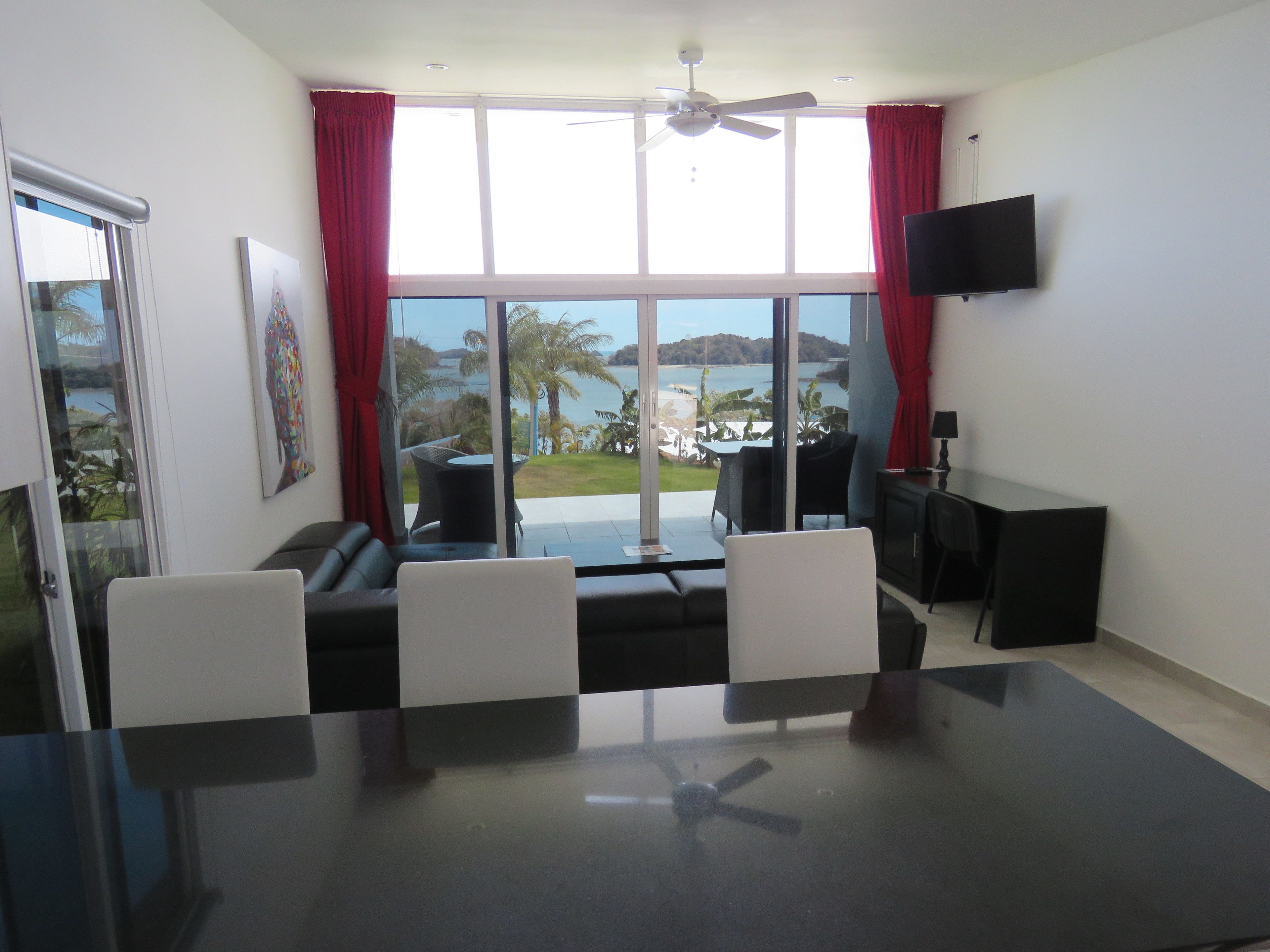 Ein modernes Zimmer mit großem Fenster, Deckenventilator, Flachbildfernseher, Esstisch und einer Couch.