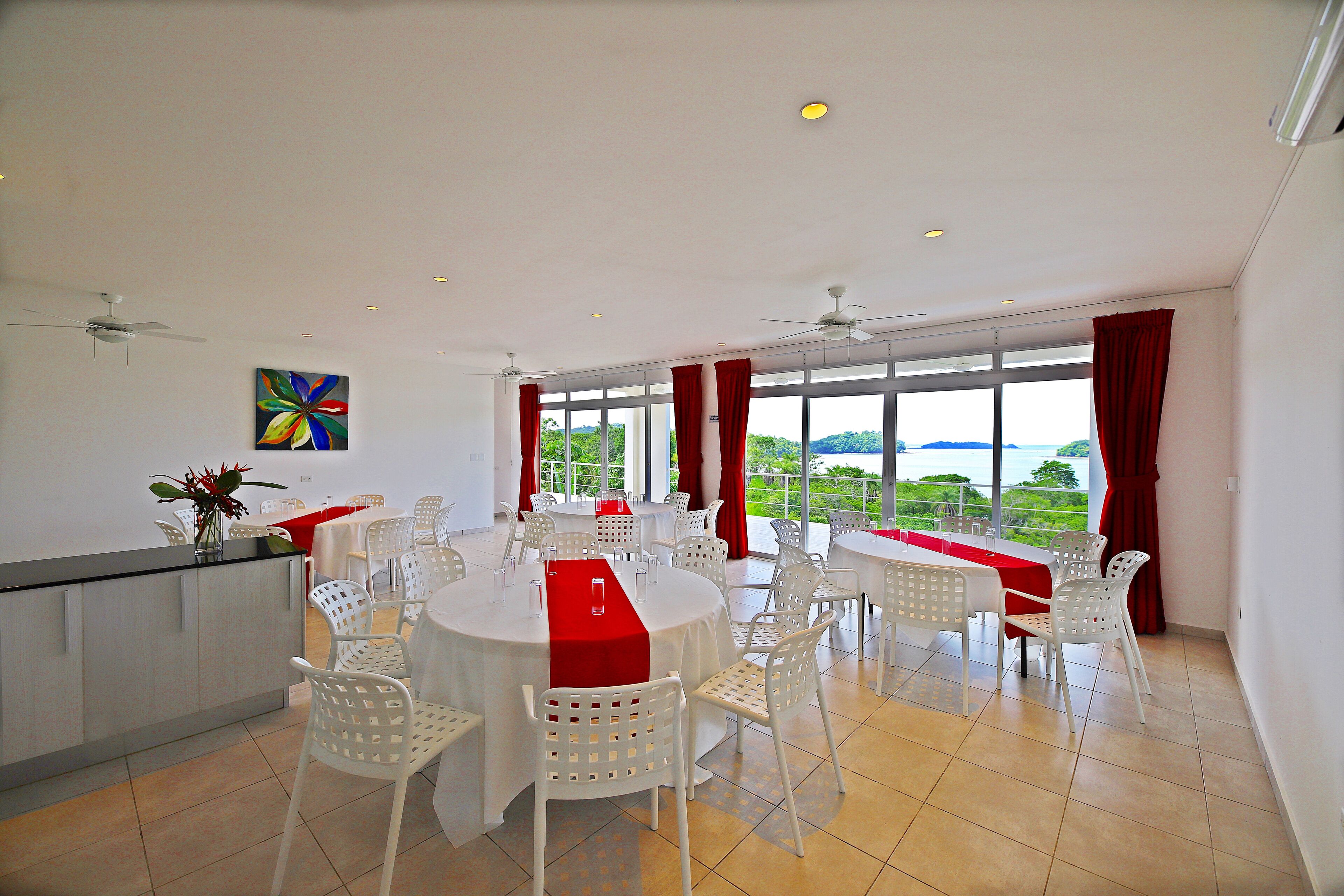 Un vaste espace dédié à la salle à manger, avec des tables rondes et des chaises blanches, de grandes fenêtres ornées de rideaux rouges et un ventilateur de plafond.