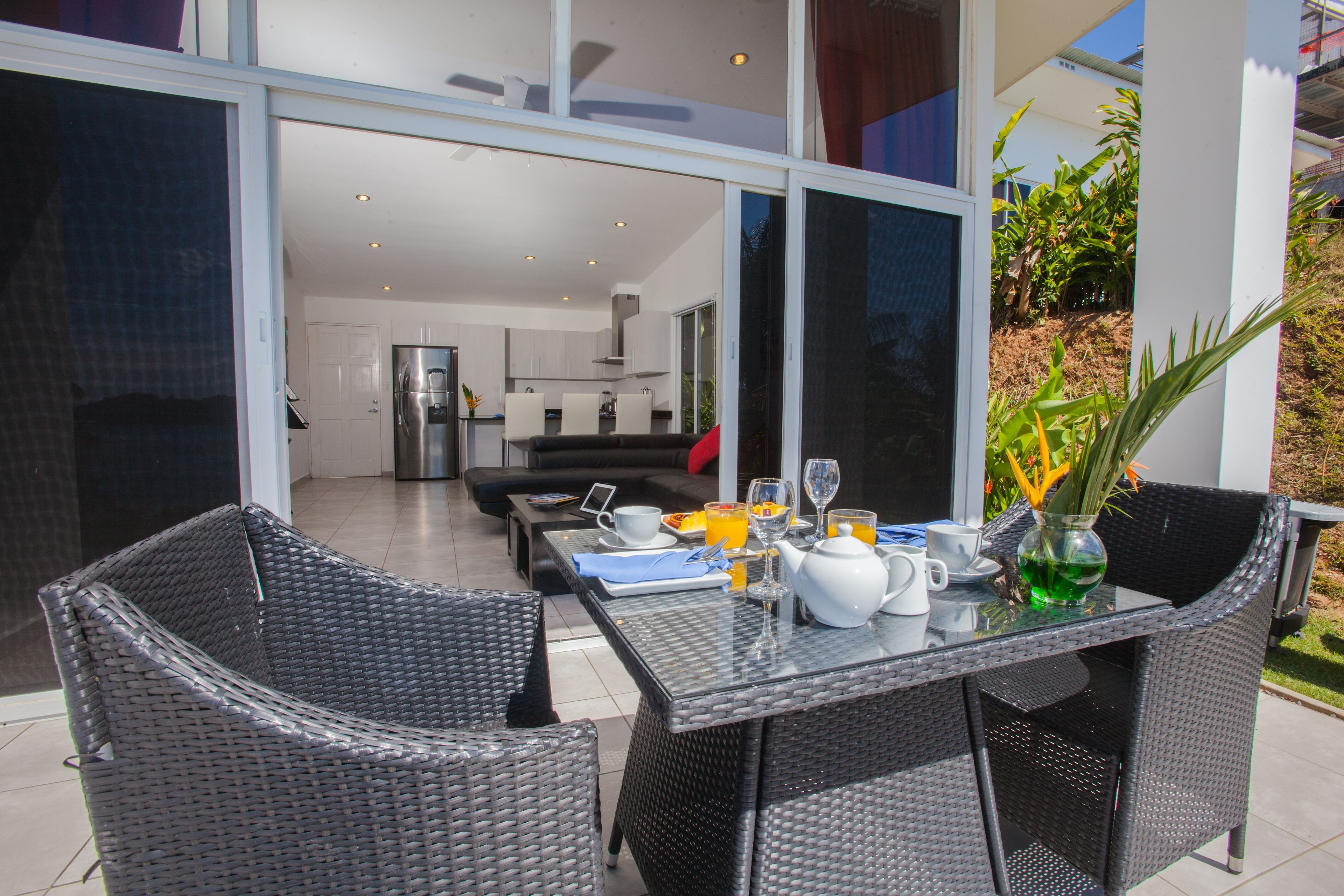 A modern outdoor dining area with wicker chairs and a glass table, featuring breakfast items and a vase of flowers.