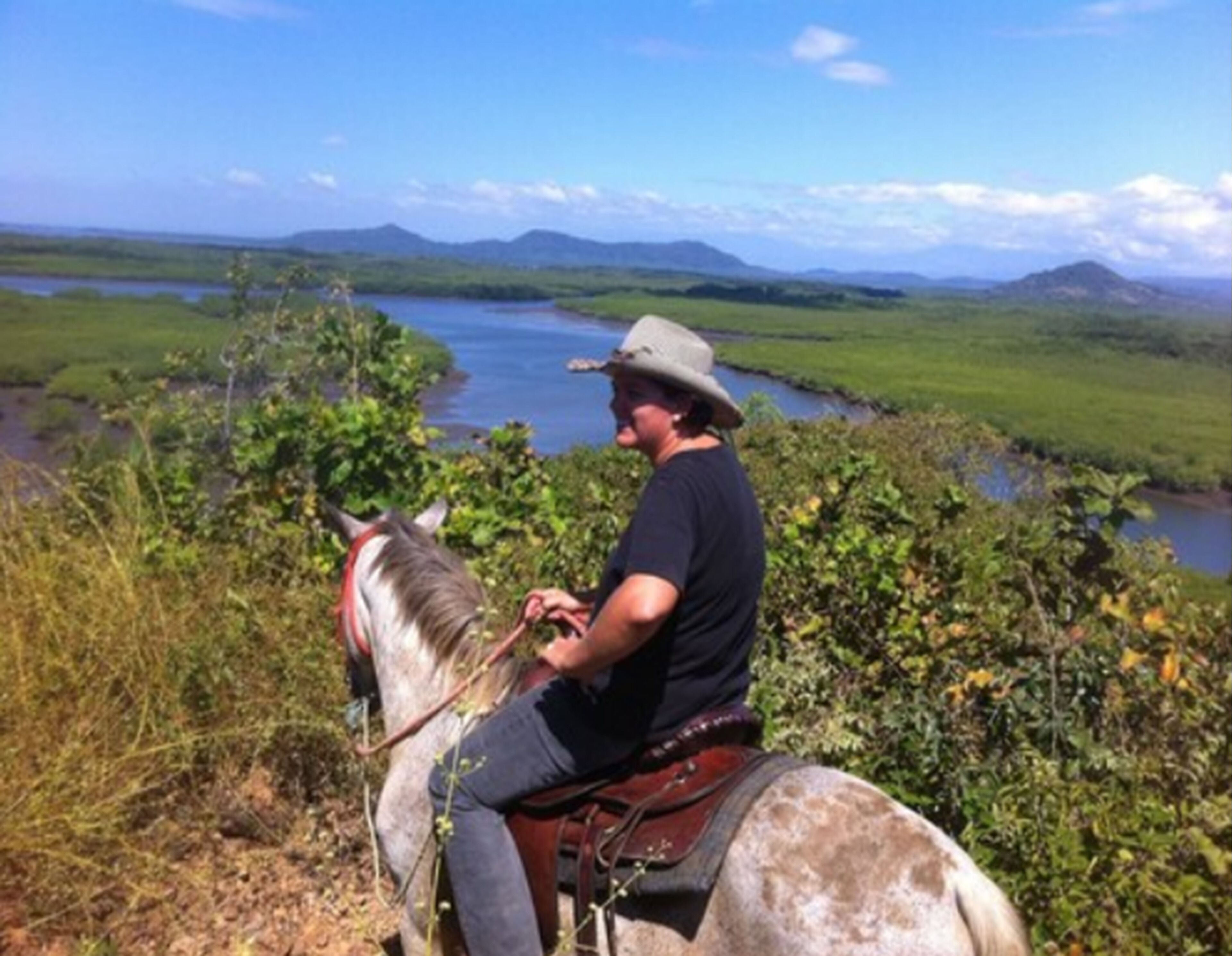 A man riding a horse on a trail with a river and hills in the background.