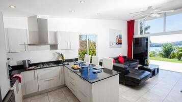 Una cocina moderna con armarios blancos, encimera negra y una isla central. Hay una zona de comedor con un sofá negro y una mesa de centro. La habitación tiene ventanas amplias con vistas a zonas verdes y un cuerpo de agua.
