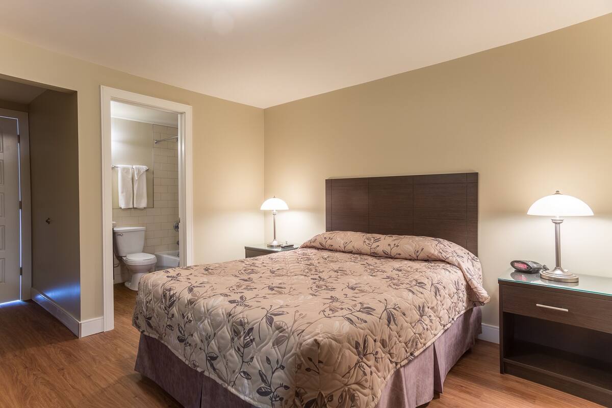 A bedroom with a bed, bedside tables, a nightstand, a headboard, and a bathroom with a toilet and towel rack.