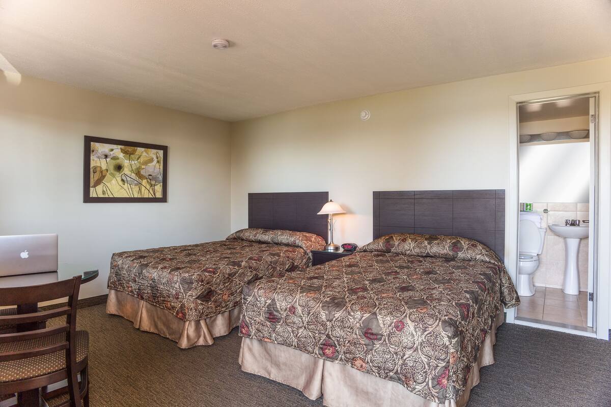 A hotel room with two beds, a desk with a laptop, a lamp, and a bathroom visible through an open door.
