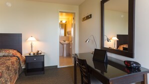 Quarto de hotel com cama, mesa de cabeceira com abajur, escrivaninha com cadeira, espelho e banheiro visível por uma porta aberta.