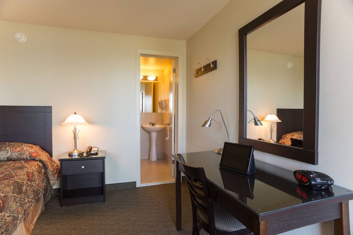 A hotel room with a bed, bedside table with a lamp, a desk with a chair, a mirror, and a bathroom visible through an open door.