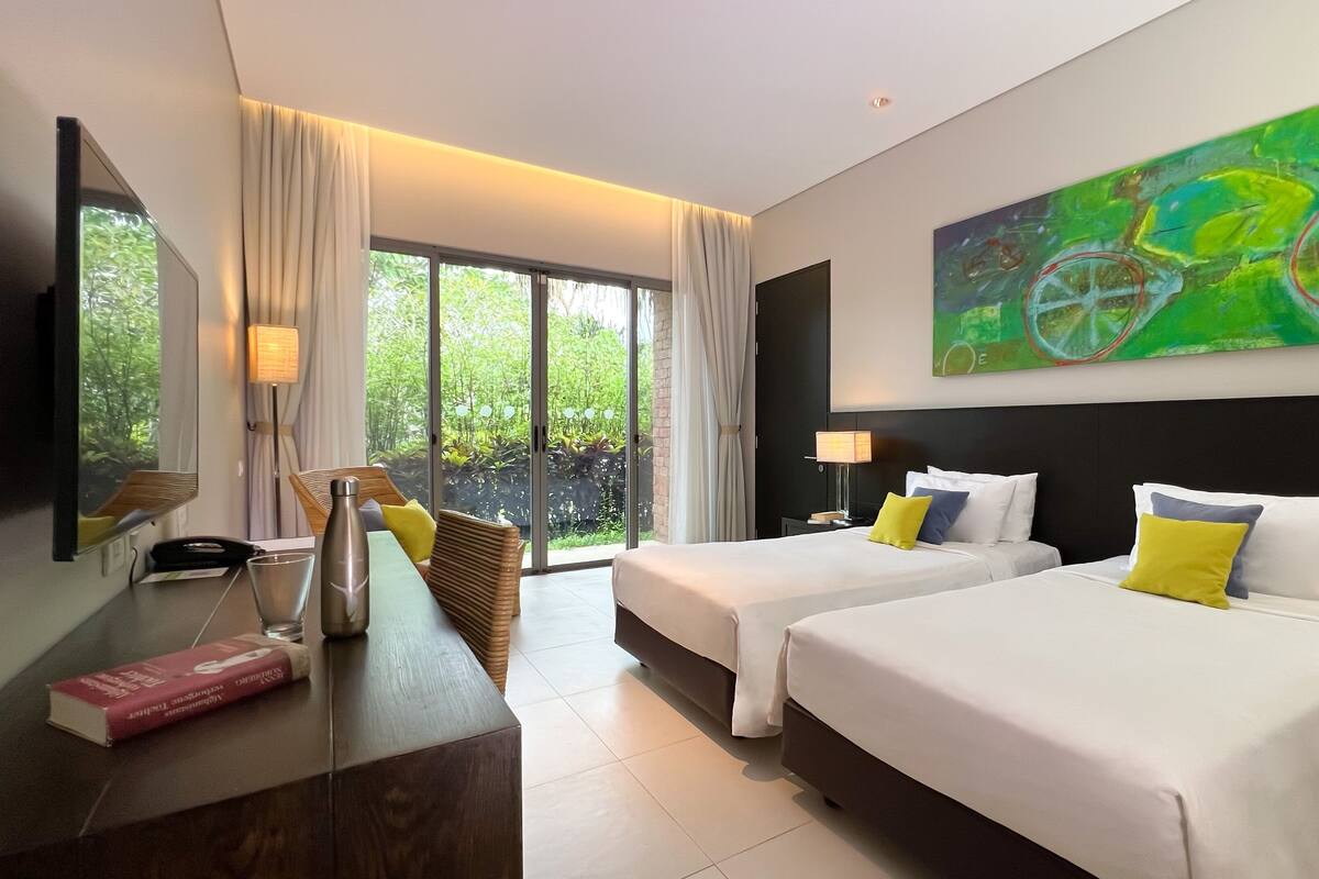 A hotel room with two beds, a desk, a chair, a large window with curtains, a painting on the wall, and a telephone.