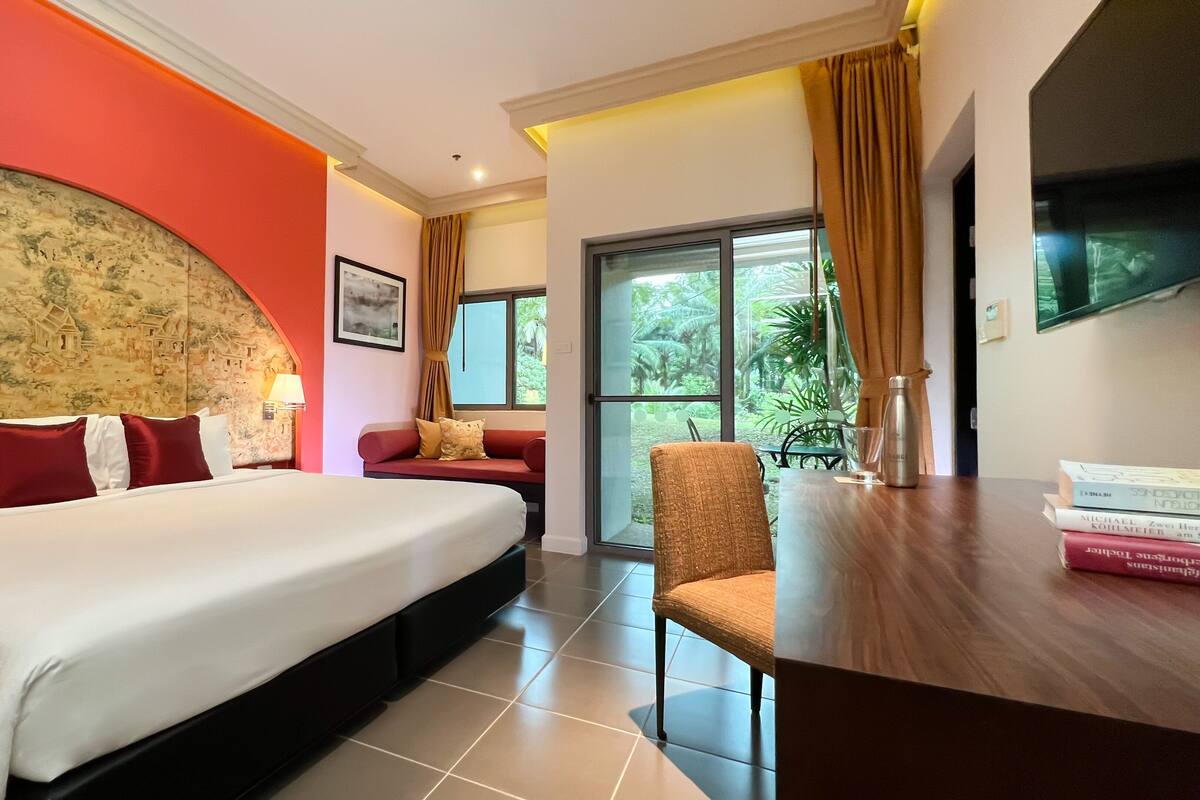 A hotel room with a large bed, a red sofa, a wooden chair, and a dining table with books.