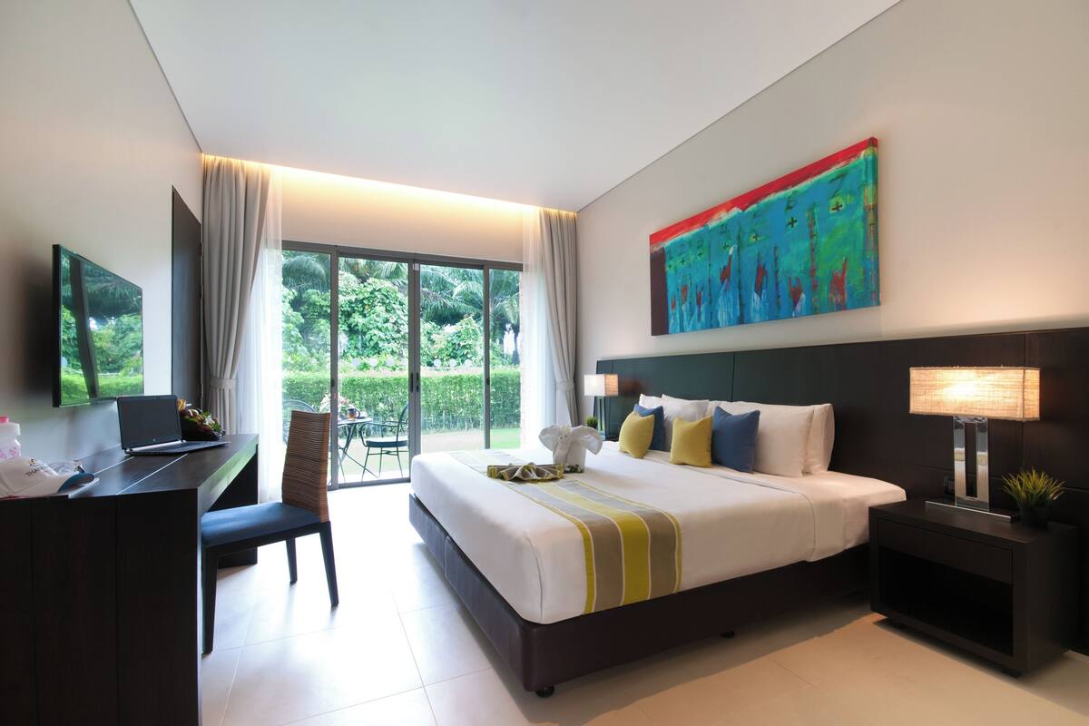A modern hotel room with a large bed, a desk, and a laptop. There is a painting on the wall and a view of greenery outside.