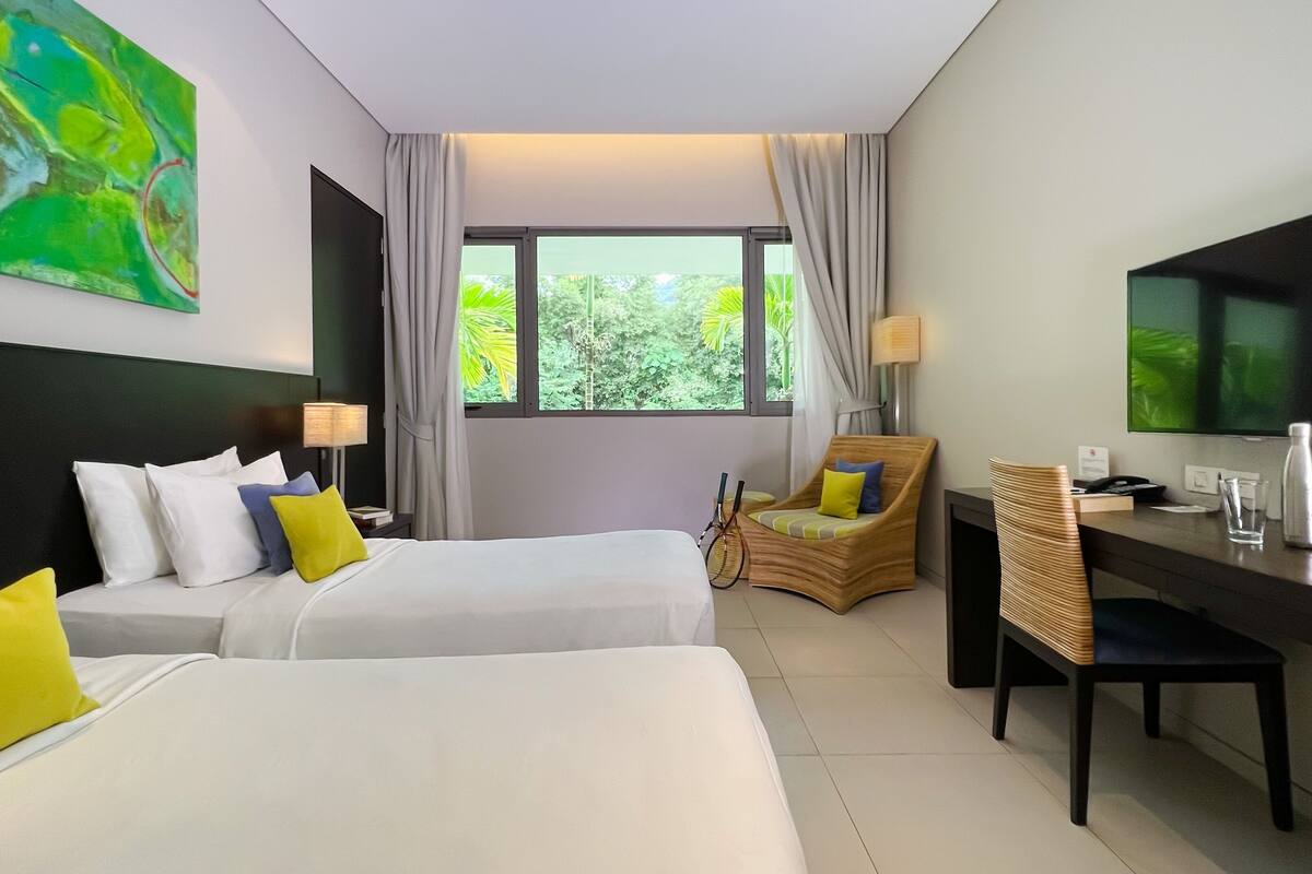 A hotel room with two beds, a desk, a chair, a TV, and a window with a view of greenery.