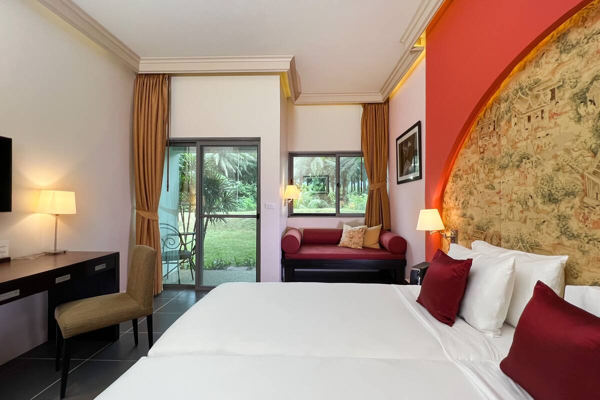 A hotel room with a bed, desk, chair, and a television. There is a view of a garden through the windows.