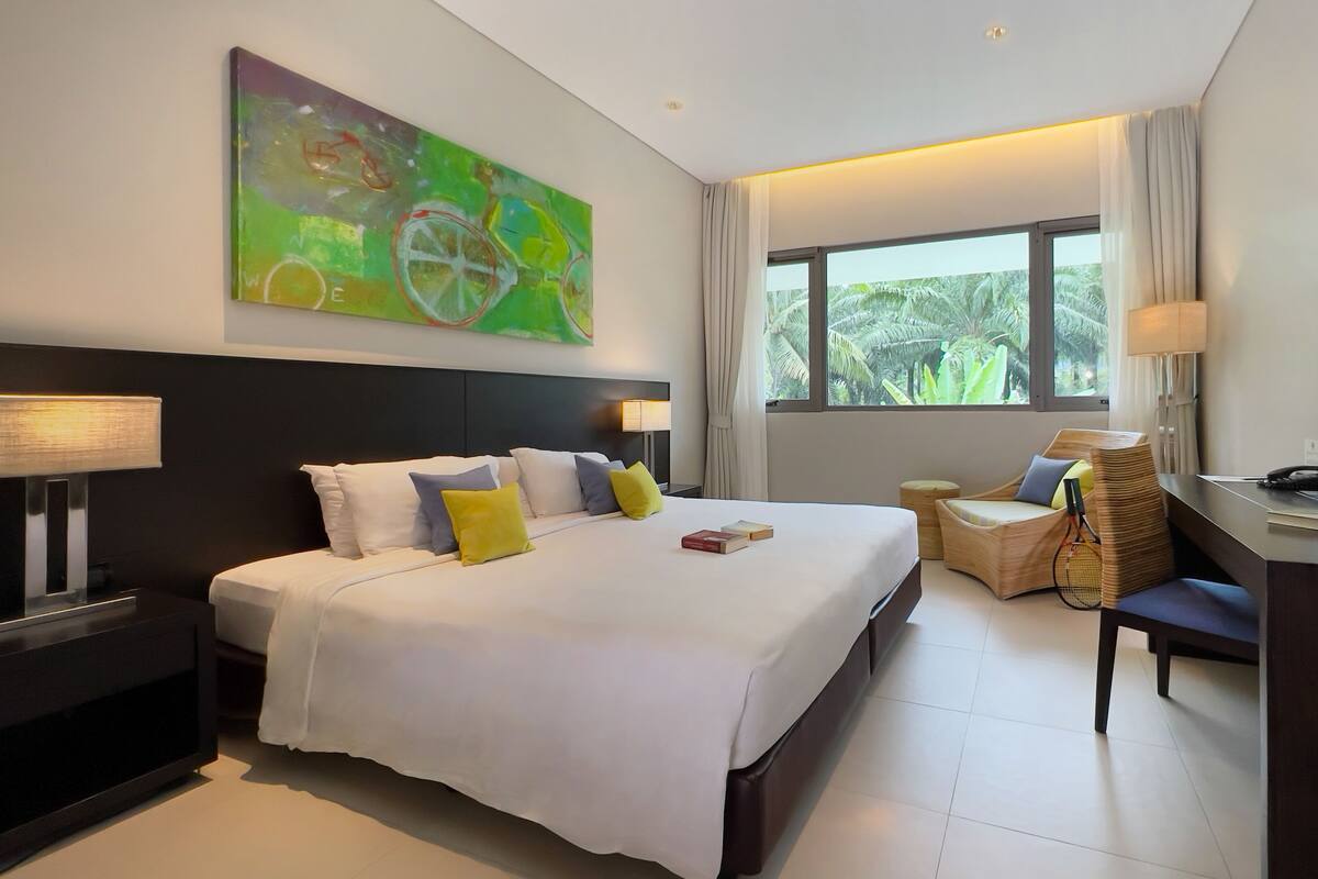A hotel room with a large bed, a desk with a chair, a television, and a window with a view of palm trees.