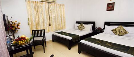 A hotel room with two single beds, a desk with a chair, a small table with a fruit bowl, and a window with curtains.