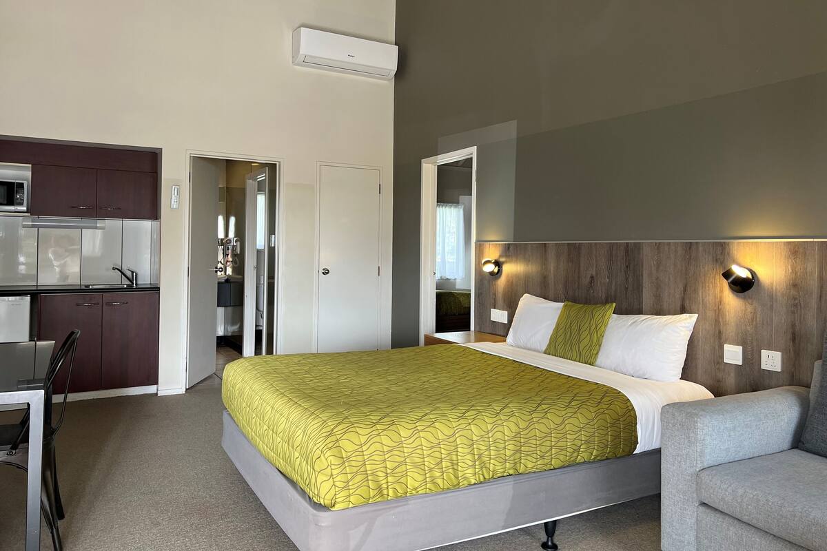 A modern hotel room with a bed, a chair, a desk, and a kitchenette.