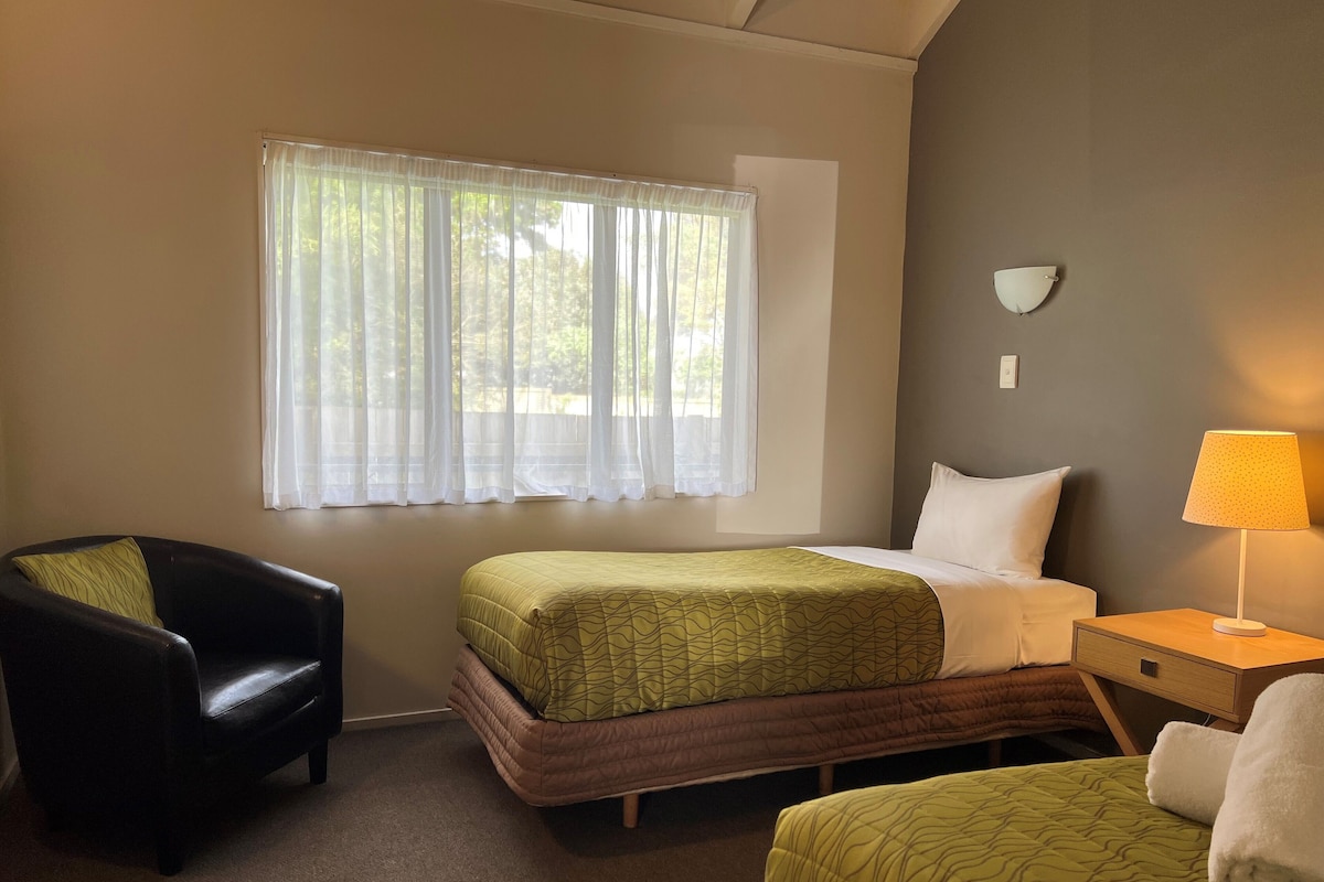 A hotel room with a bed, a chair, a nightstand with a lamp, and a window with sheer curtains.