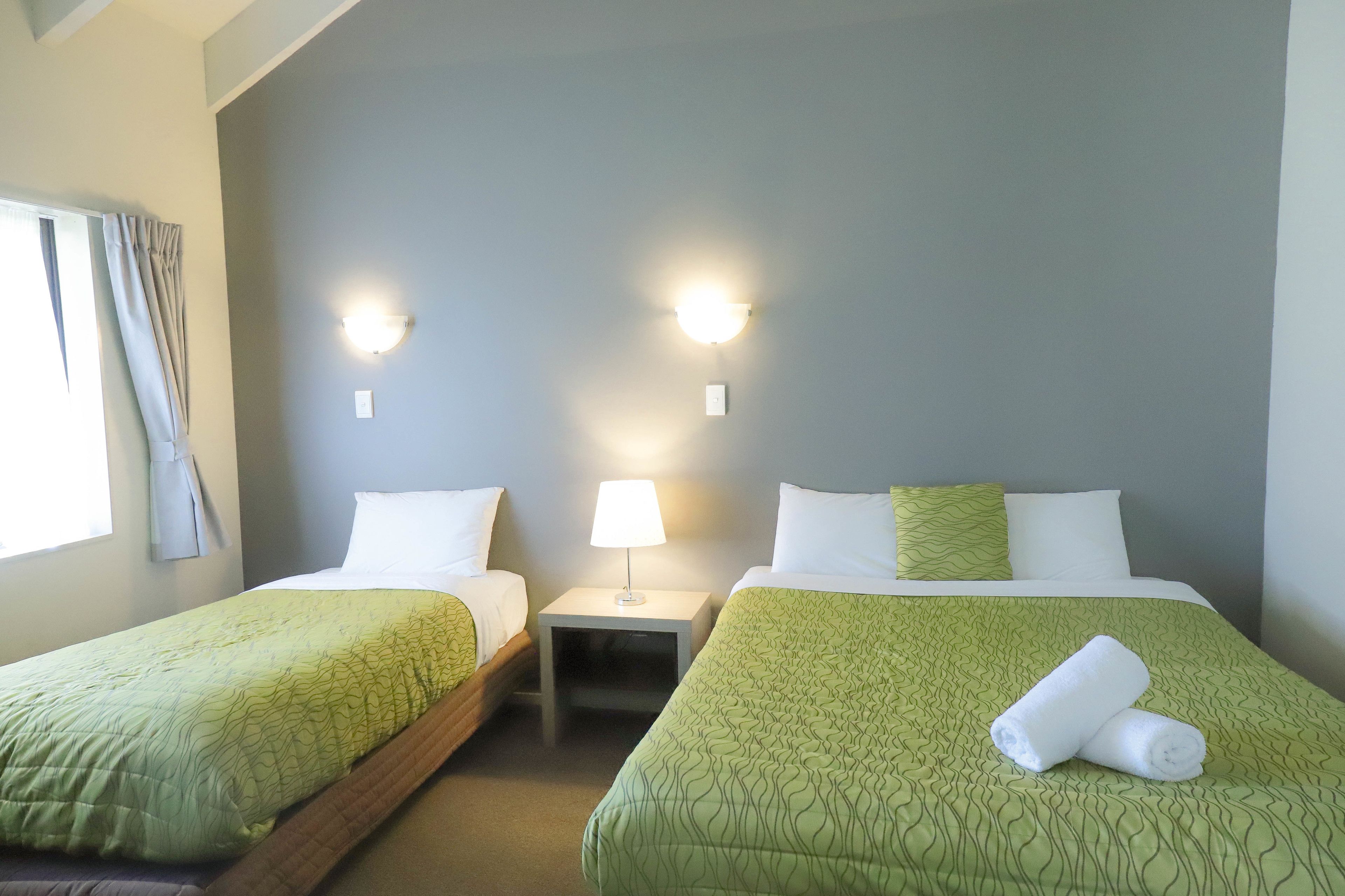 A hotel room with two single beds, each with a green patterned bedspread and white pillows. There is a nightstand with a lamp between the beds, and a window with curtains on the left side.