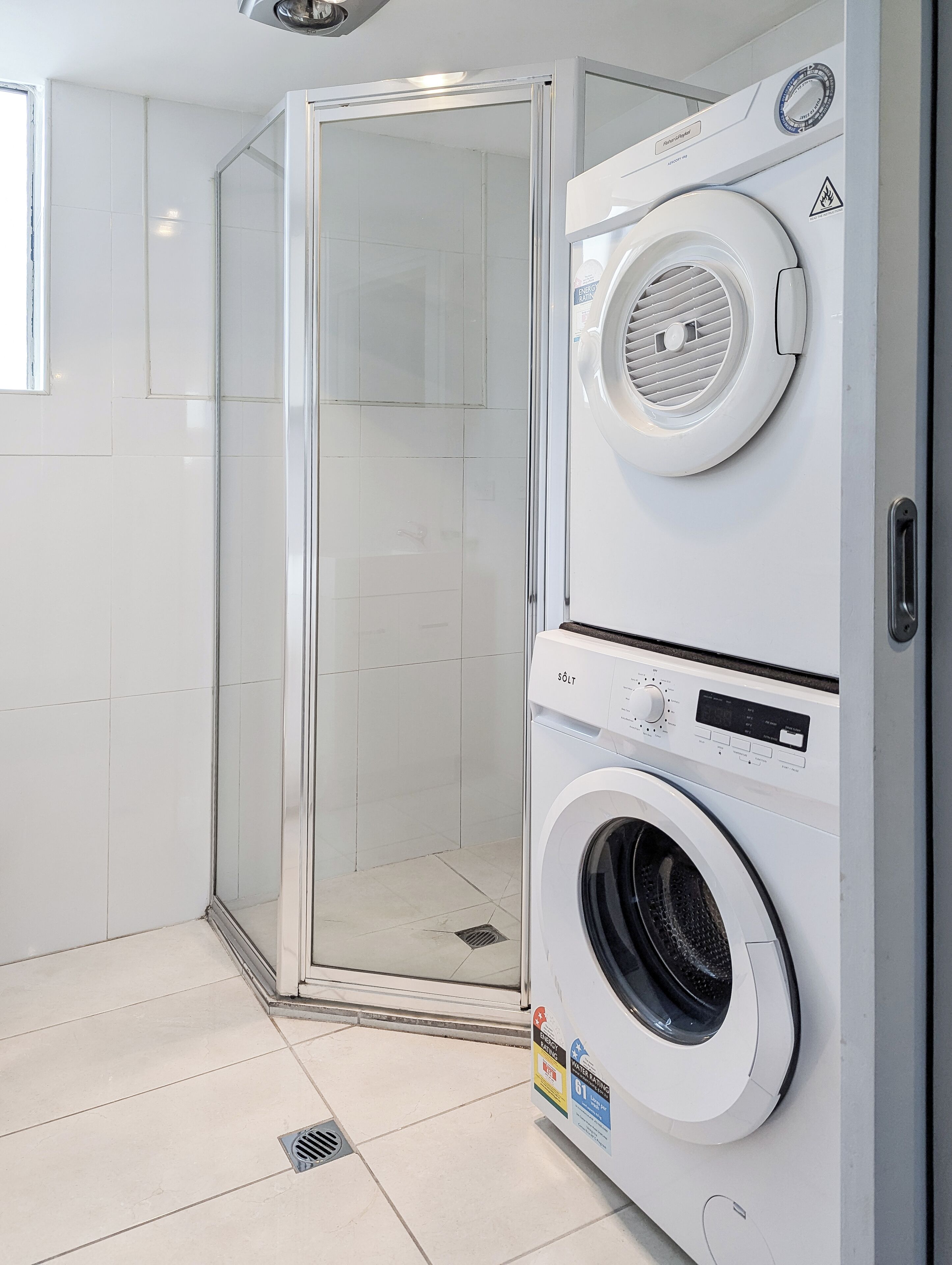 Un lavadero moderno con lavadora y secadora de carga frontal, cabina de ducha con mampara de vidrio y piso de baldosas.