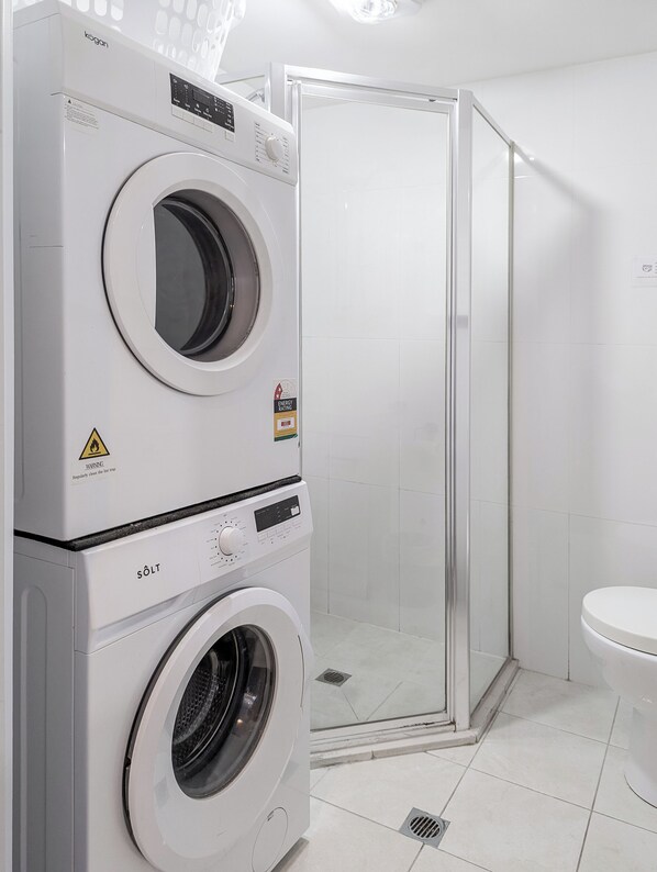 A compact laundry area with a front-loading washer and dryer, a glass shower enclosure, and a white toilet.