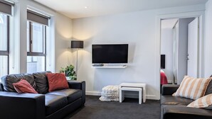 A modern living room with a black leather sofa, a small white side table, a flat-screen TV mounted on the wall, and a large window with blinds. - Apartments on Chapman (North Melbourne)