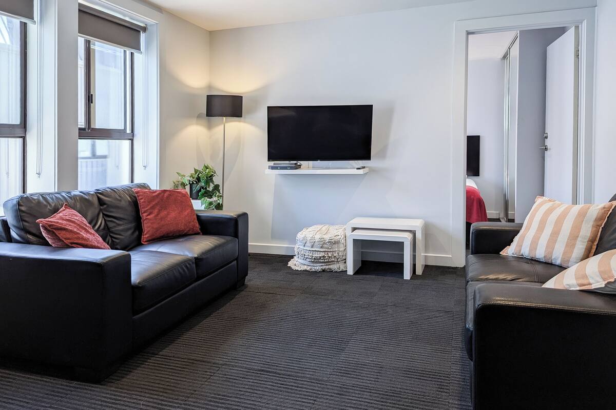 Ein modernes Wohnzimmer mit einer schwarzen Ledersofa, einem kleinen weißen Beistelltisch, einem flachbildfernseher an der Wand und einem großen Fenster mit Jalousien.