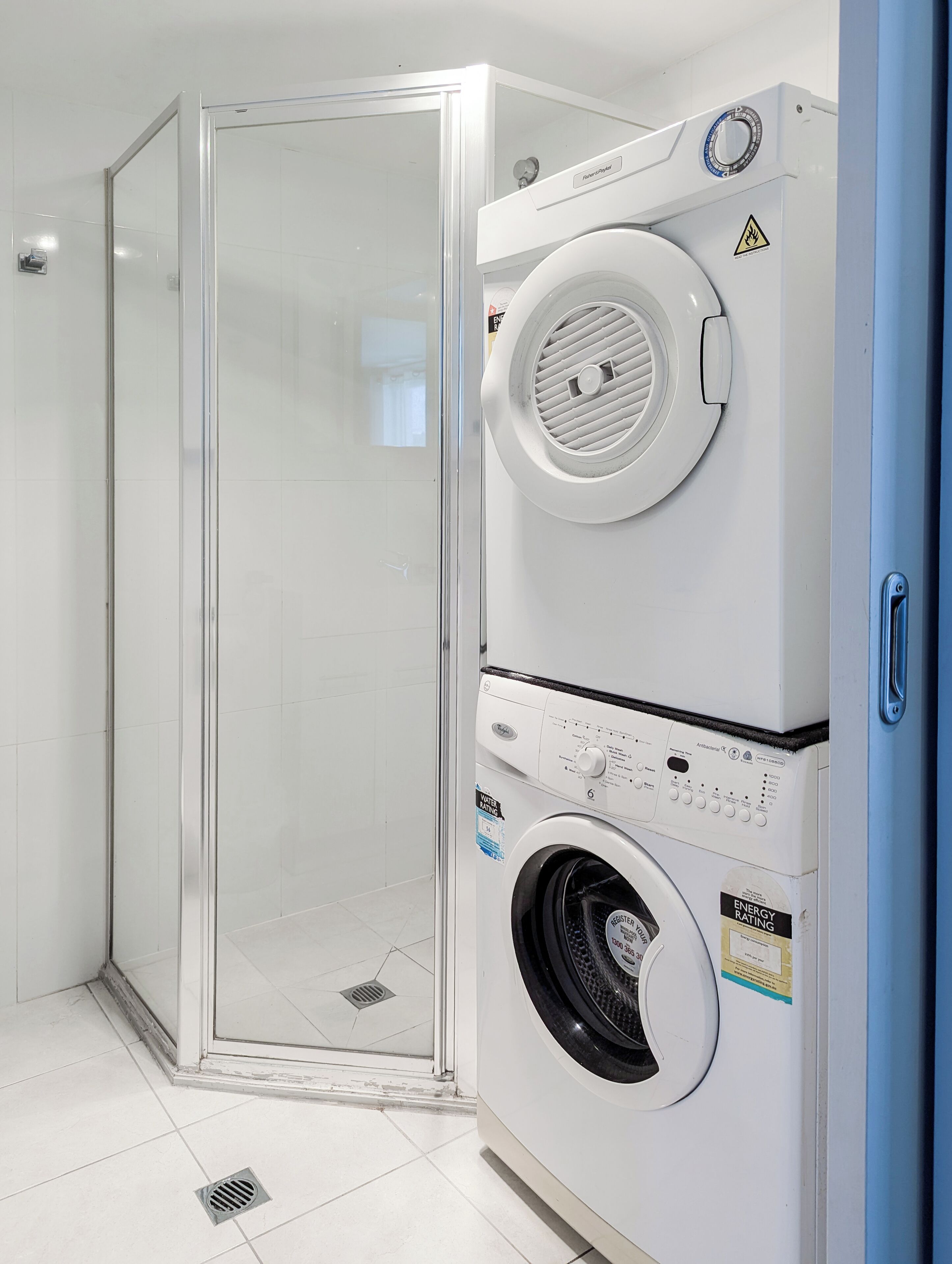 Ein moderner Waschraum mit einer gläsernen Dusche, einem gestapelten Wasch- und Trocknerpaar und einer blauen Tür.