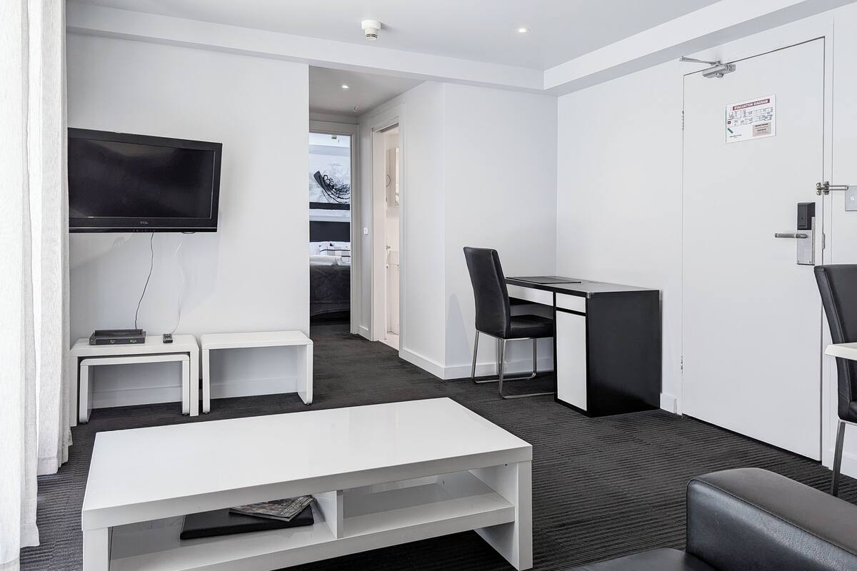Ein modernes Hotelzimmer mit weißem Schreibtisch, schwarzem Bürostuhl, einem an der Wand montierten Flachbildfernseher, einem kleinen weißen Nachttisch mit Fernbedienung, einer weißen Couch und einem schwarzen Ledersessel.