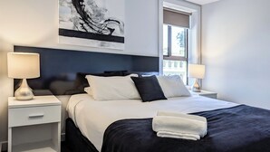 A modern hotel room with a large bed, two bedside tables, a nightstand, a window with a view, and a piece of abstract art on the wall. - Apartments on Chapman (North Melbourne)