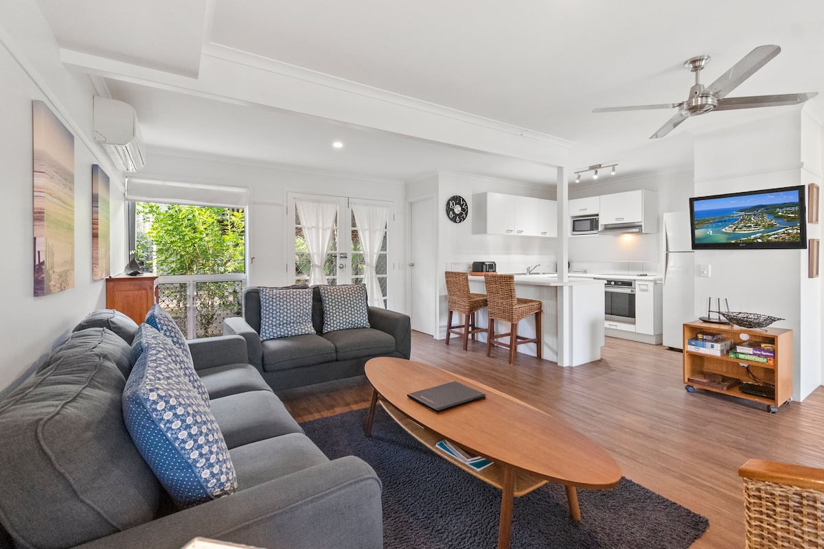 A modern living room with a sofa, coffee table, and a dining area with chairs. There is a kitchen area with a microwave and a television.