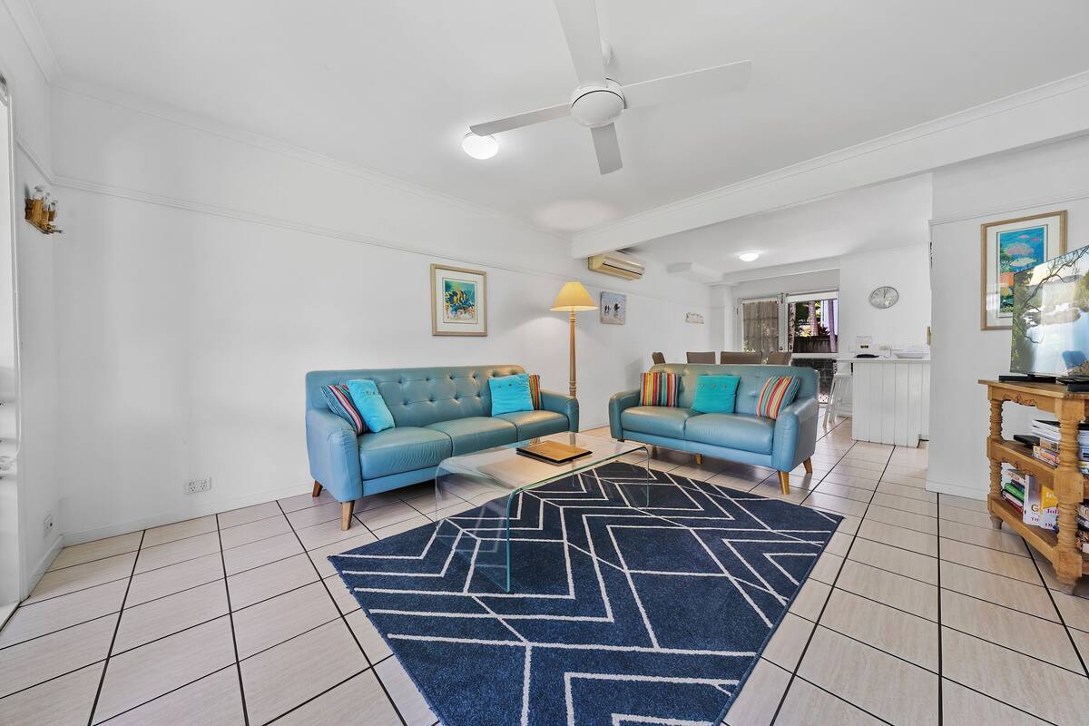 A living room with two blue sofas, a glass coffee table, a patterned rug, a ceiling fan, and a kitchen in the background.