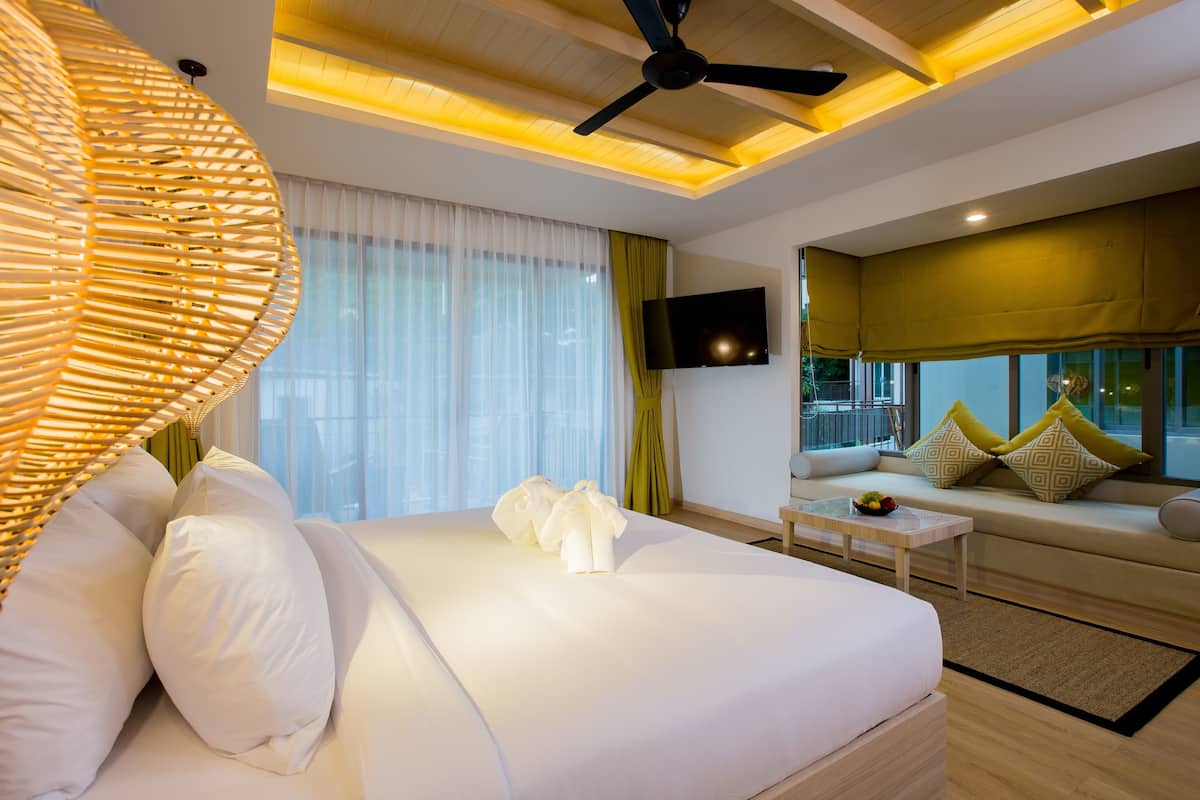 A modern hotel room with a large bed, a ceiling fan, a flat-screen TV, and a sofa with cushions.