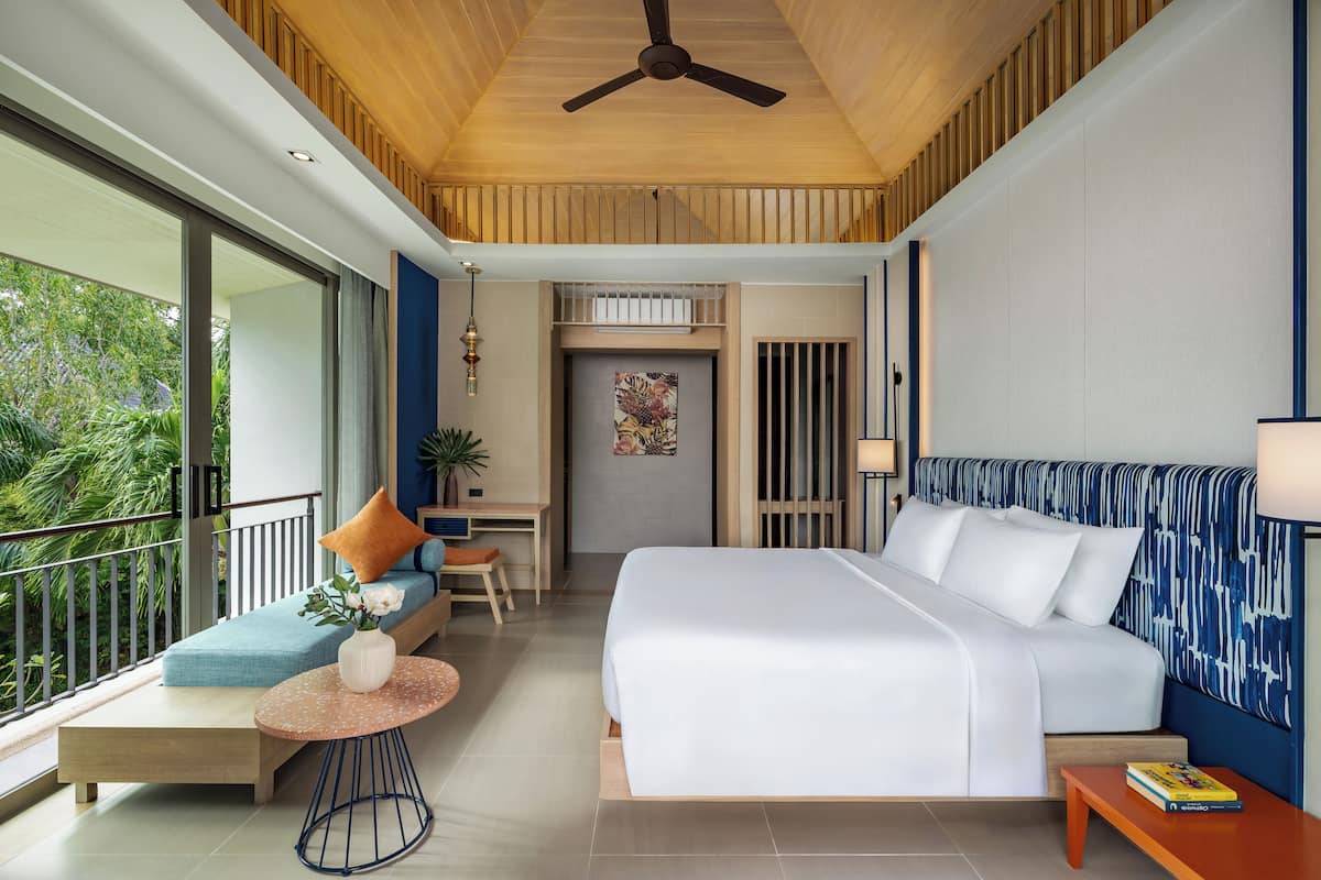 A modern hotel room with a balcony, a bed with white linens, a small seating area with a table, and a view of greenery outside.