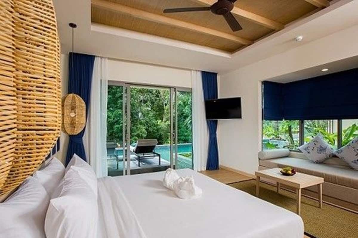 A modern hotel room with a large bed, a small table, a TV, and a view of a pool and greenery outside.