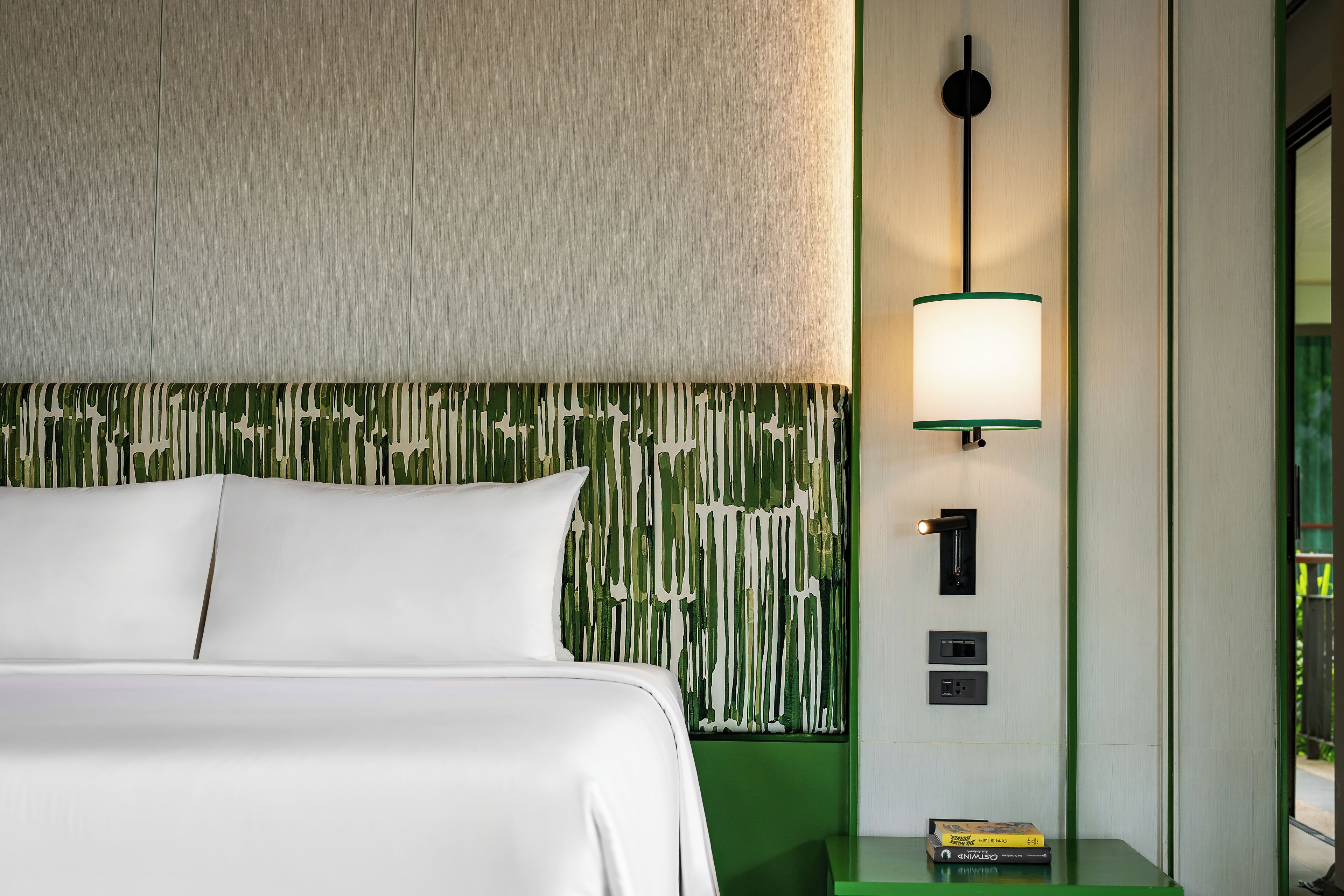 A hotel room with a bed, a green nightstand, a wall-mounted lamp, and a door with a handle.