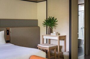 A hotel room with a bed, a small table with a plant, a chair, and a shower area visible through an open door.
