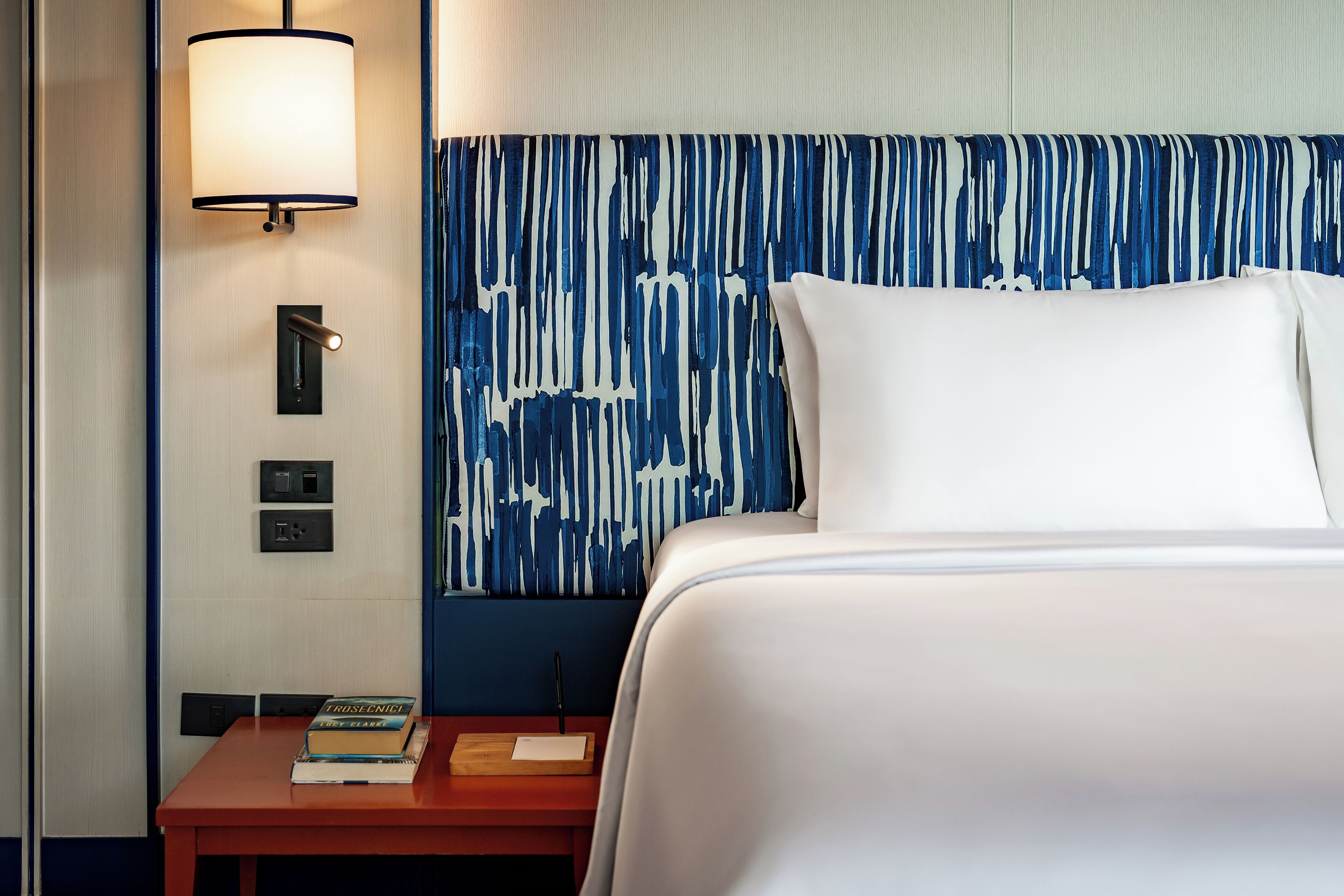 Ein Hotelzimmer mit Bett, Nachttisch mit Büchern, Lampe und Wand mit blau-weißem abstrakten Muster.