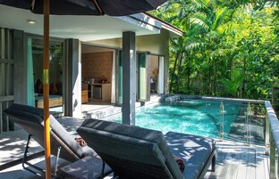 Un área junto a la piscina con sillones de descanso, una cocina con paredes de vidrio y abundante vegetación.