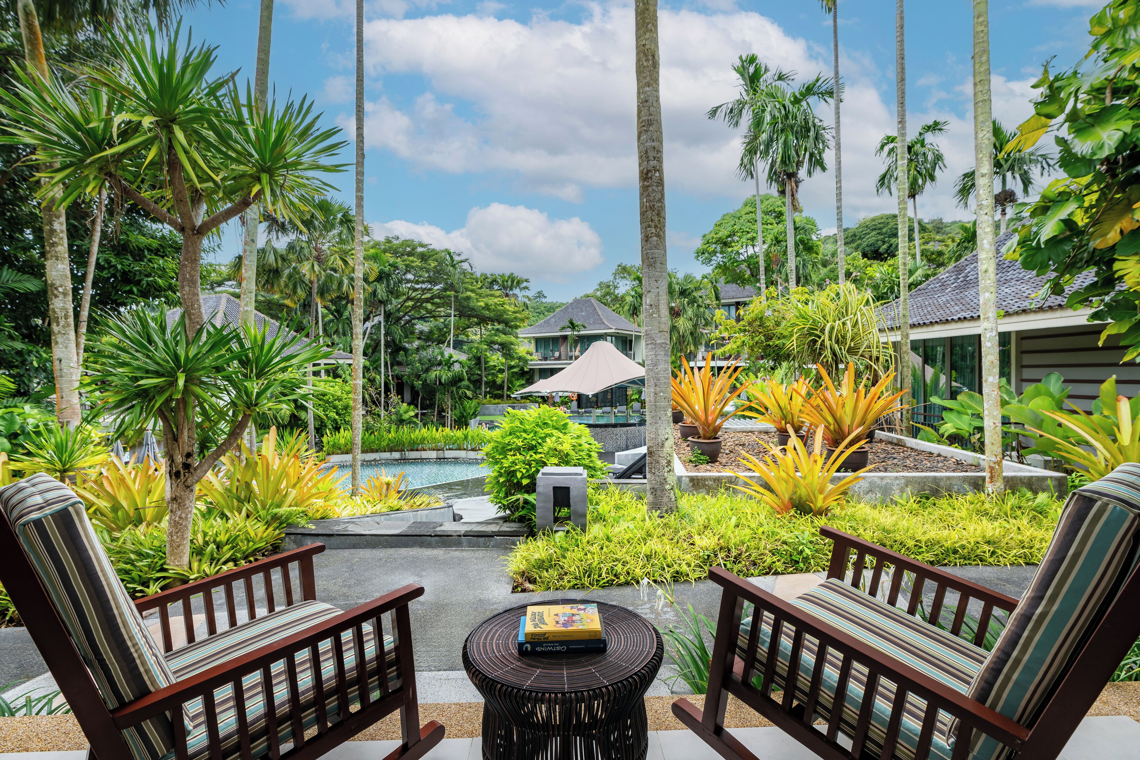 Un entorno de resort tropical con piscina, abundante vegetación y dos sillas de madera con cojines.