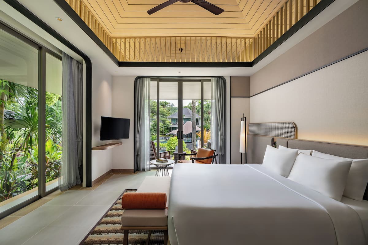 A modern hotel room with a large bed, a TV, a desk, and a balcony with a view of greenery.