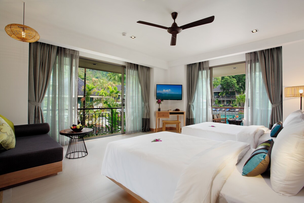 Habitación de hotel con dos camas, un escritorio, televisión y balcón con vistas al exterior.