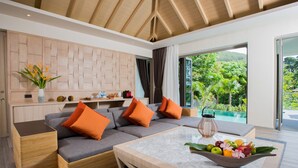 Un salon moderne avec un canapé gris, des coussins orange, une table basse en marbre agrémentée de fruits, et une vue sur une piscine et un espace verdoyant.