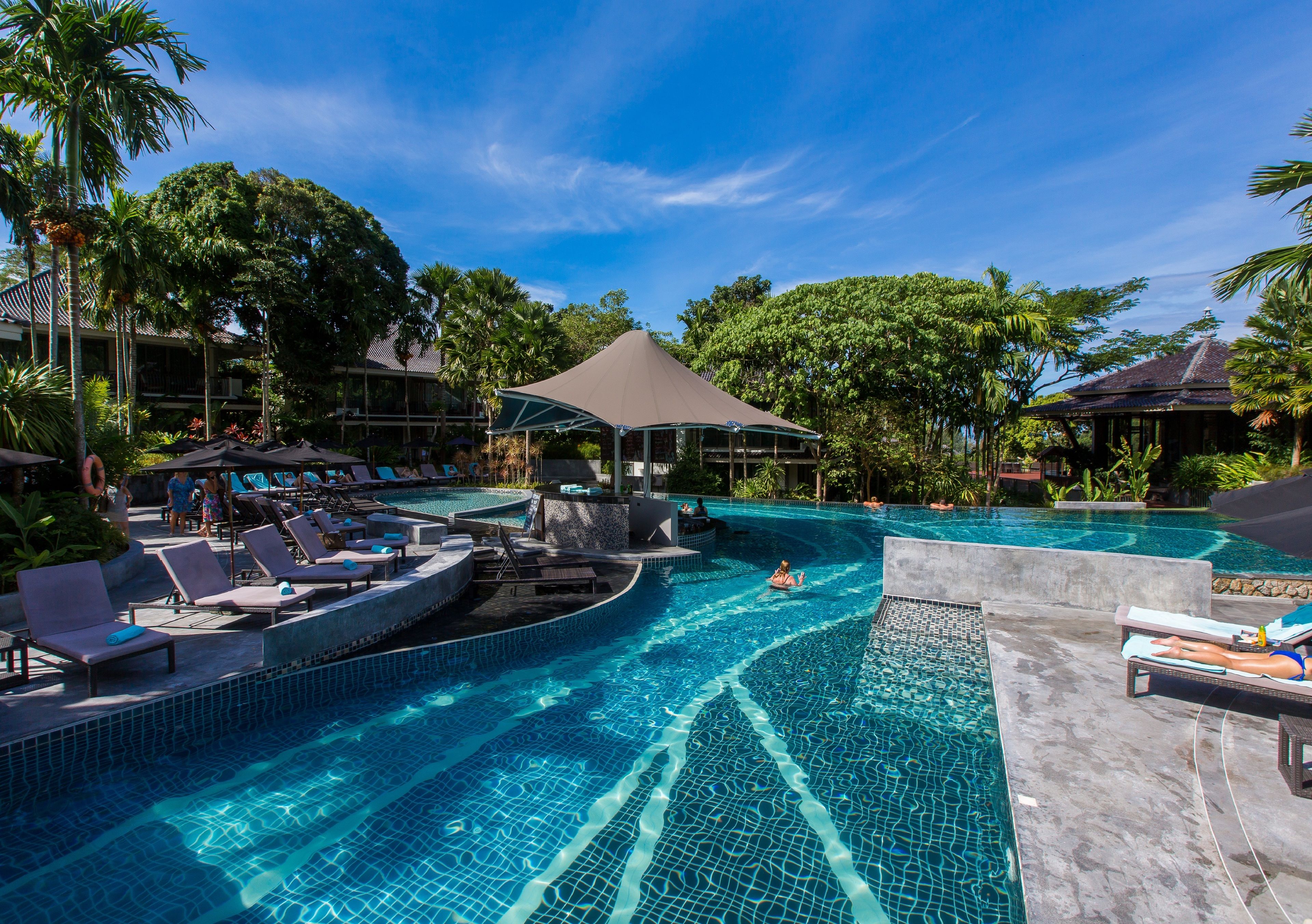 A resort pool area with lounge chairs, a covered seating area, and a clear blue sky.