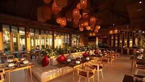 Un vaste espace dédié à la salle à manger, avec de nombreuses tables et chaises, de grandes fenêtres offrant une vue sur l’extérieur et des luminaires suspendus en fibres tressées.