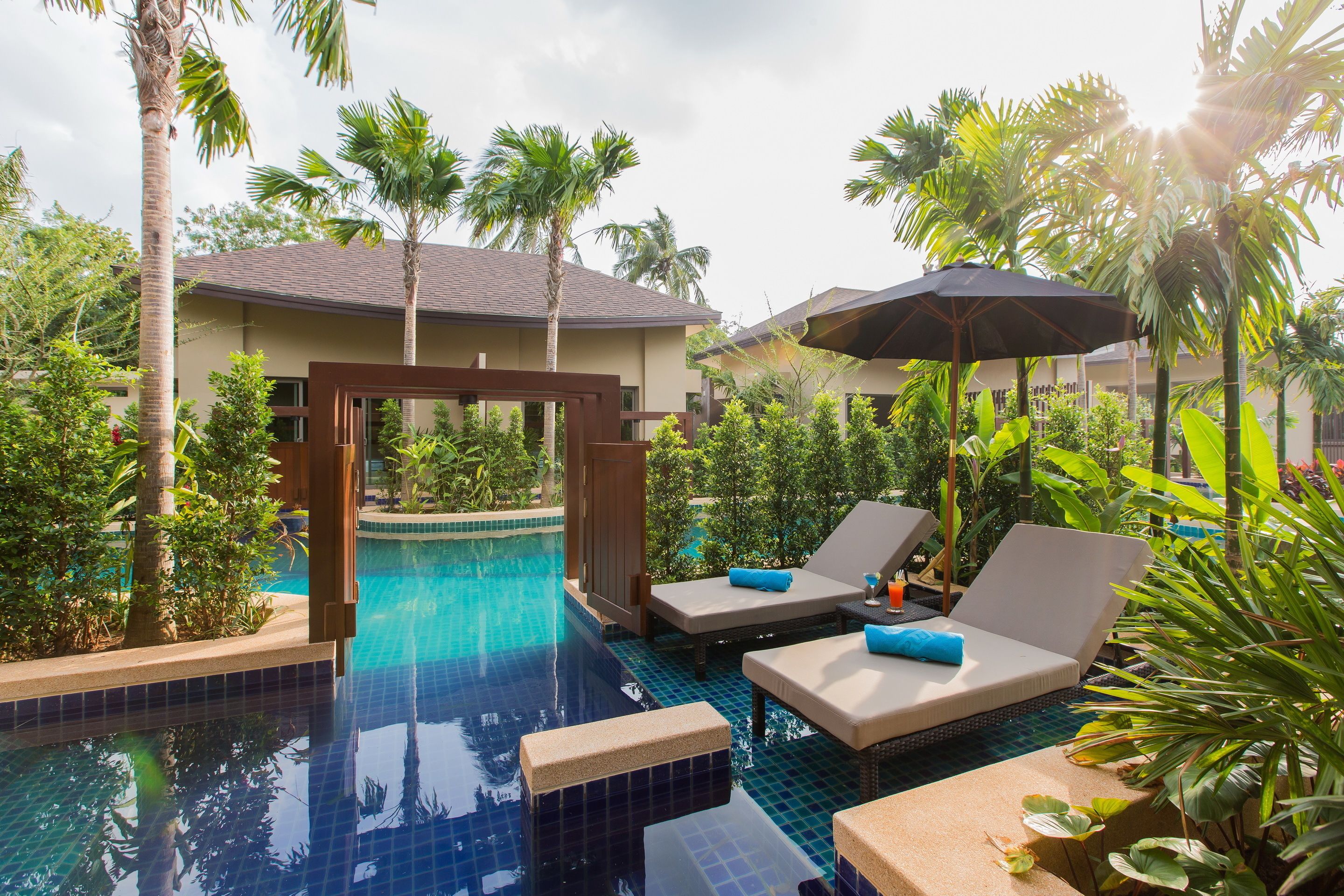 A pool area with lounge chairs and umbrellas, surrounded by palm trees and greenery.