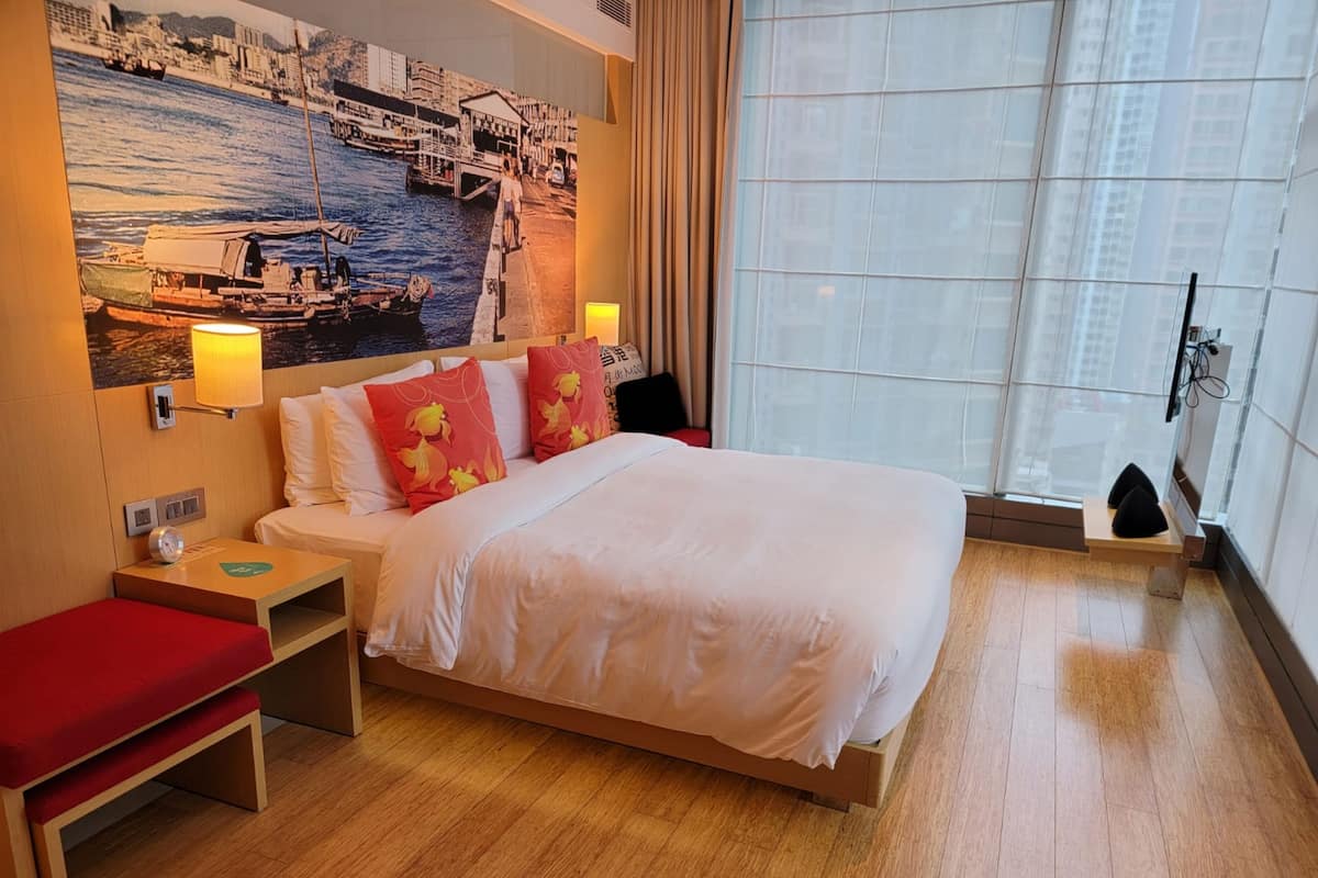 Quarto de hotel com cama, mesas de cabeceira, um banco vermelho, uma janela grande e um mural com vista para a cidade na parede.